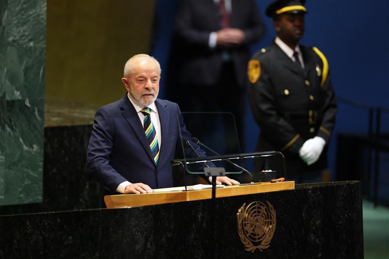 Lula, homem branco de cabelo e barba brancos, com terno azul e gravata verde e amarela, discursando em palanque com símbolo da ONU.
