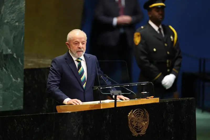 Lula, homem branco de cabelo e barba brancos, com terno azul e gravata verde e amarela, discursando em palanque com símbolo da ONU.