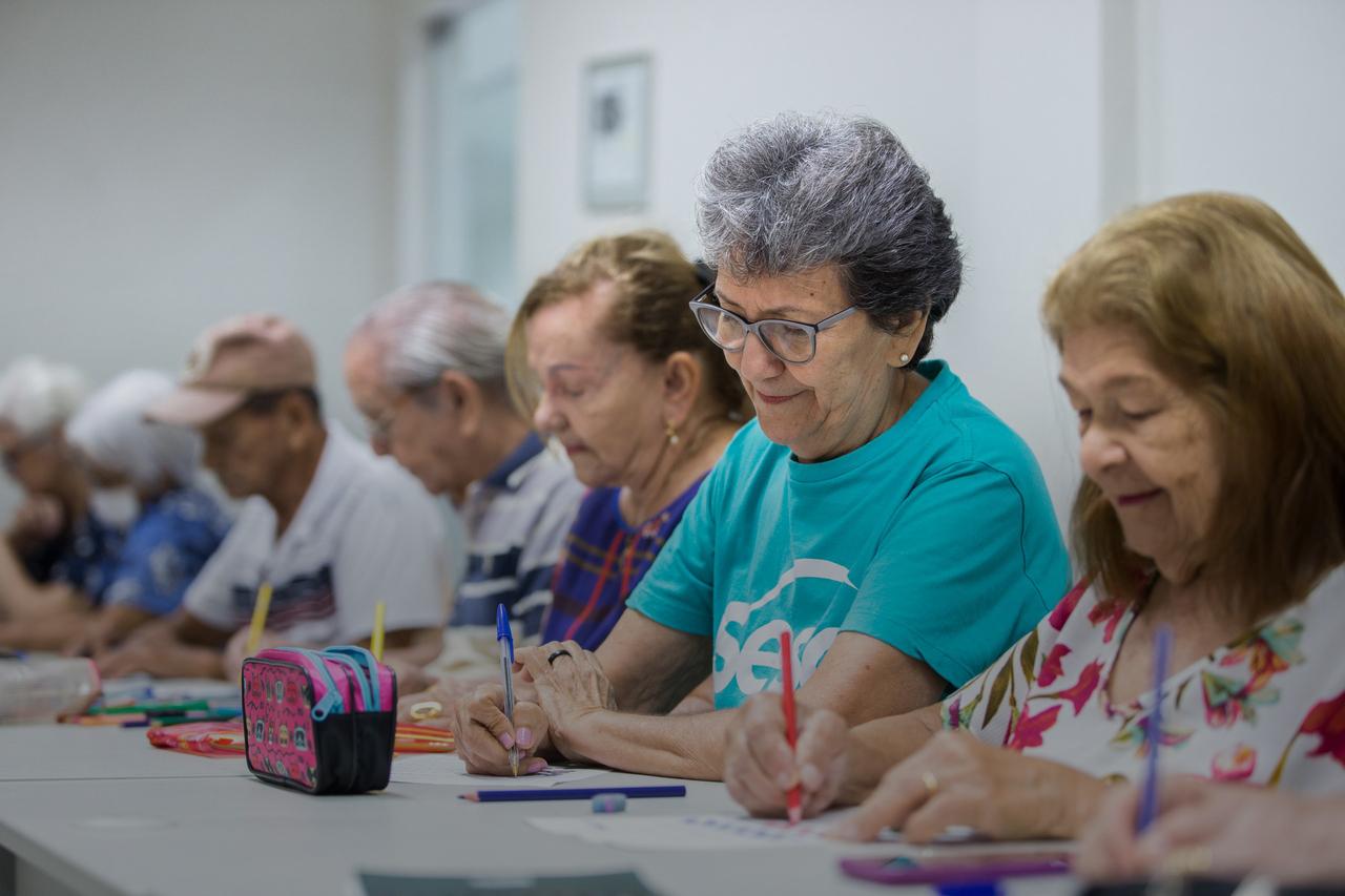 Imagem mostra pessoas idosas sentadas à mesa, com canetas nas mãos e escrevendo em um papel.
