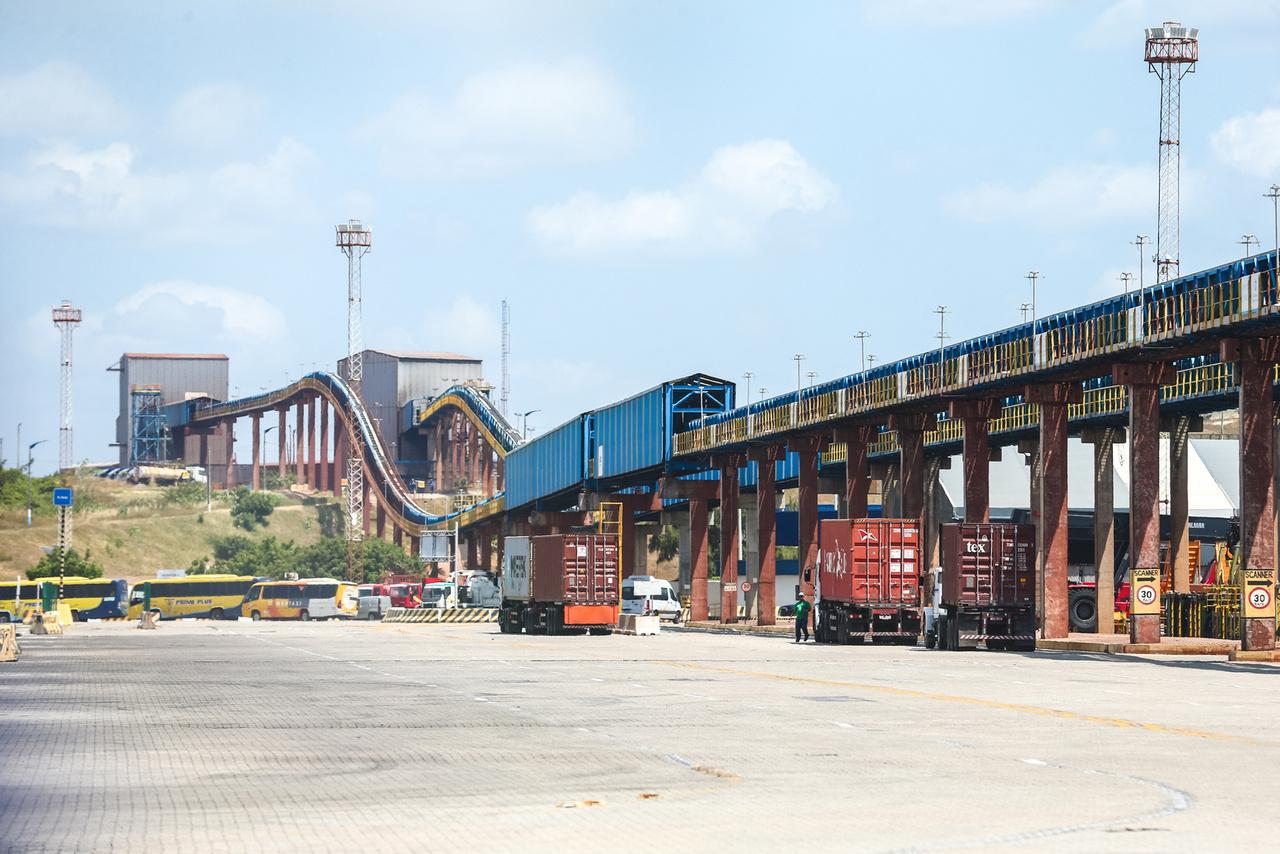 Imagem em plano aberto mostra esteiras de cargas no Complexo Industrial e Portuário do Pecém. Também aparece um caminhão à direita e um ao centro da imagem, além de alguns ônibus à esquerda.
