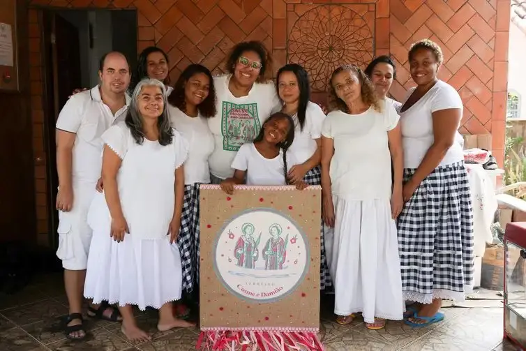 A imagem mostra um grupo de pessoas vestidas de branco e saias quadriculadas, reunidas em frente a um painel decorado com a imagem de São Cosme e São Damião. Elas sorriem para a foto em clima de celebração e devoção.