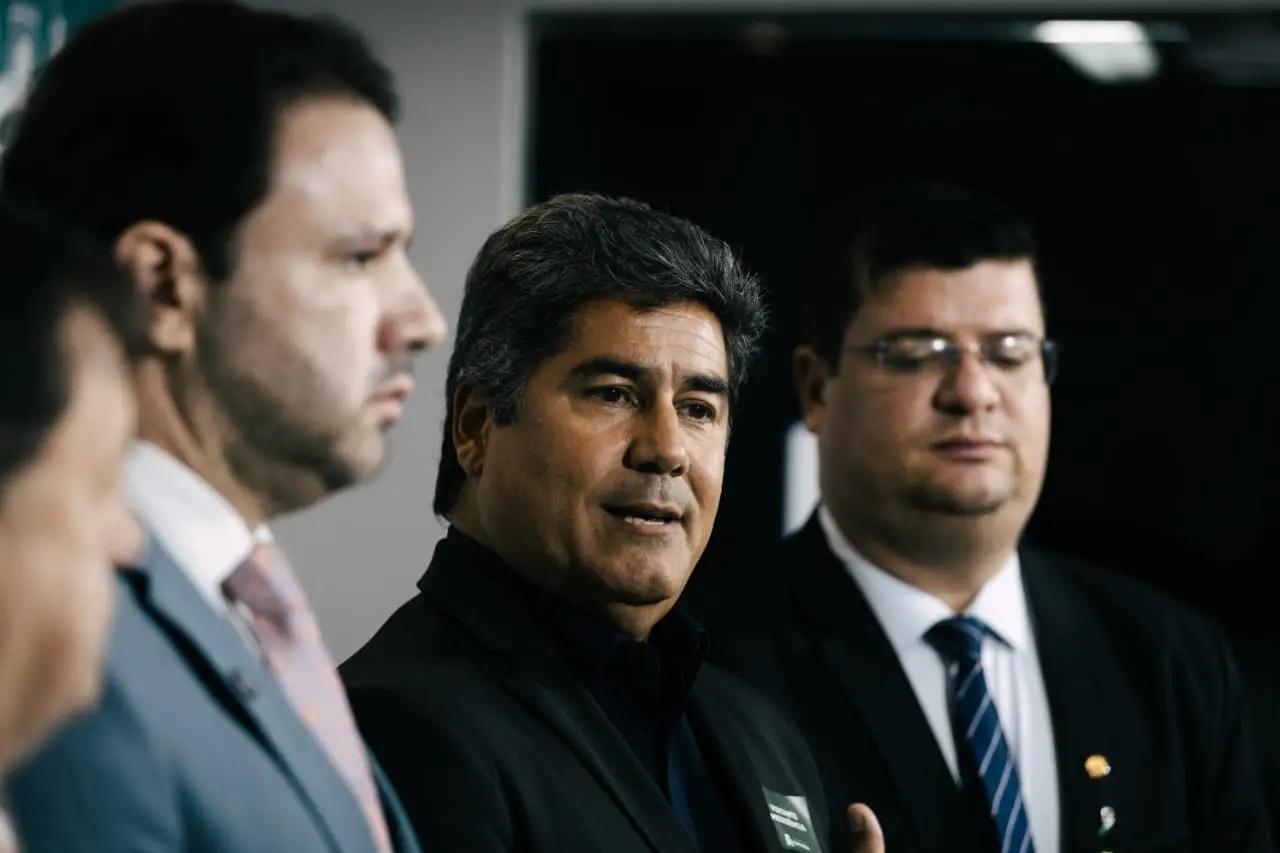 Da esquerda para a direita, em coletiva de imprensa: Léo Couto, presidente da Câmara; Márcio Cardeal, secretário das Finanças; e Bruno Mesquita, líder do Governo Evandro na Câmara. A imagem mostra três homens vestidos com ternos e gravatas em um ambiente formal. O homem ao centro, de cabelos grisalhos e expressão séria, está falando, enquanto os outros dois, à esquerda e à direita, ouvem atentamente. O fundo é neutro, sugerindo um espaço interno institucional.