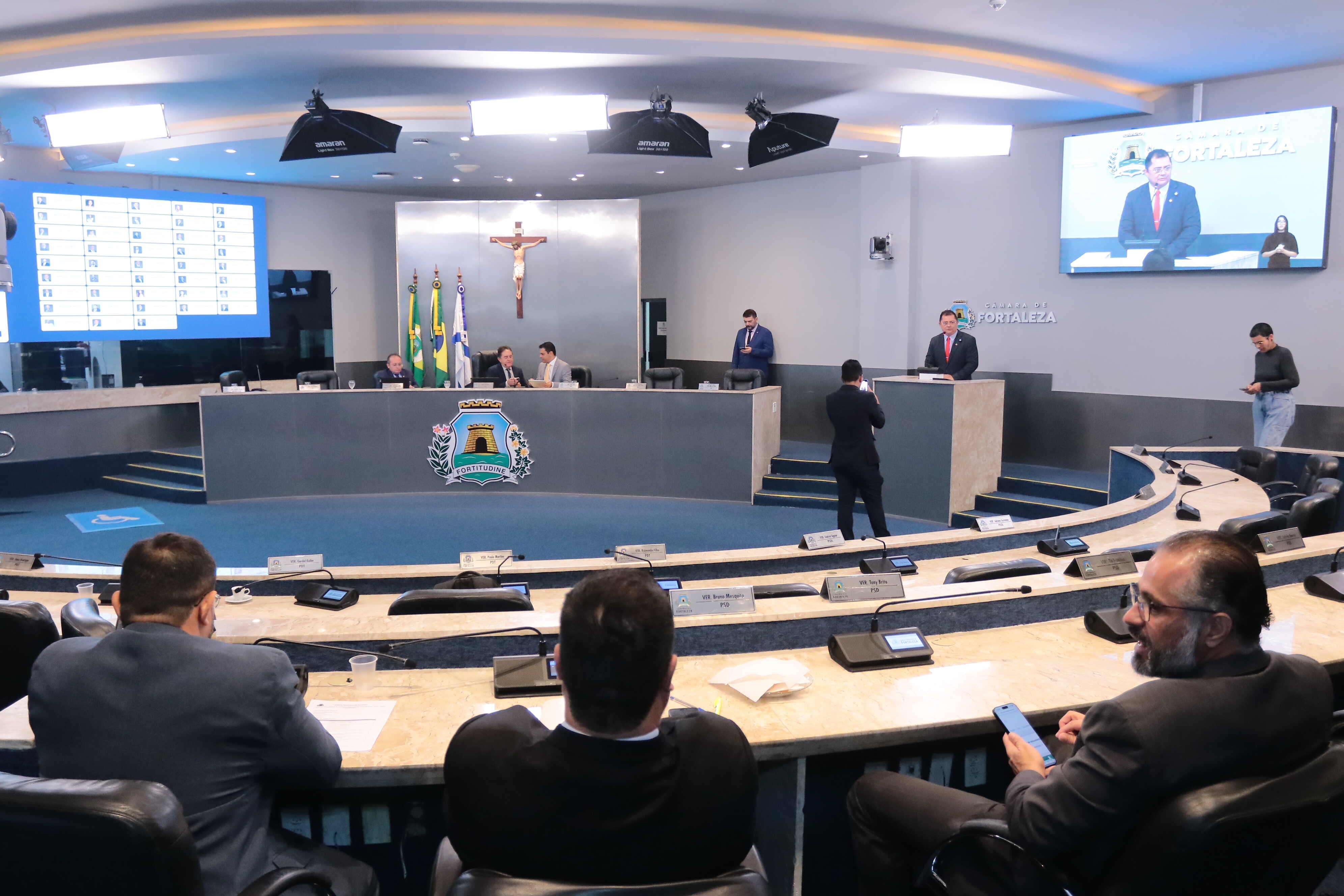Imagem mostra o plenário da Câmara Municipal de Fortaleza durante uma sessão. Ao centro, está a mesa diretora com três pessoas sentadas, atrás de um painel com o brasão da cidade e ao fundo bandeiras do Brasil, do Ceará e de Fortaleza, além de um crucifixo na parede. À direita, um vereador fala na tribuna, enquanto sua imagem aparece em um telão. Outros parlamentares e assessores estão distribuídos pelo espaço, alguns sentados e outros em pé, com computadores e papéis sobre as bancadas em formato semicircular. O ambiente é iluminado por refletores de teto e possui dois painéis eletrônicos exibindo informações da sessão.