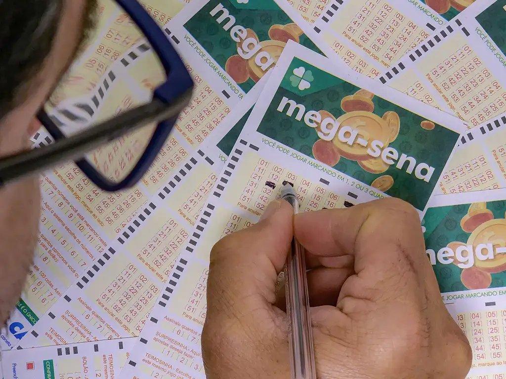 Imagem mostra pessoa marcando números da aposta na cartela de loteria Mega-Sena, cercada por várias apostas e usando um óculos de leitura, com foco na atividade de marcar o bilhete da loteria federal.