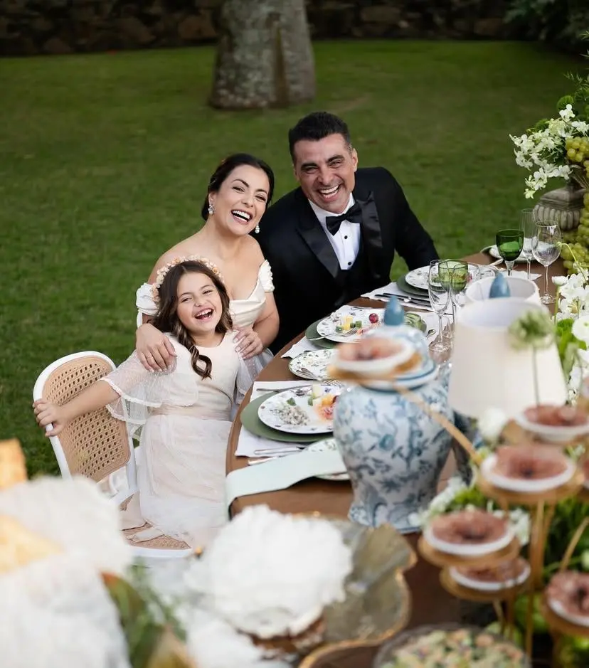 Família de JP Mantovani comemorando uma ocasião especial ao ar livre, com uma mesa decorada com pratos, flores e doces, demonstrando alegria e união.