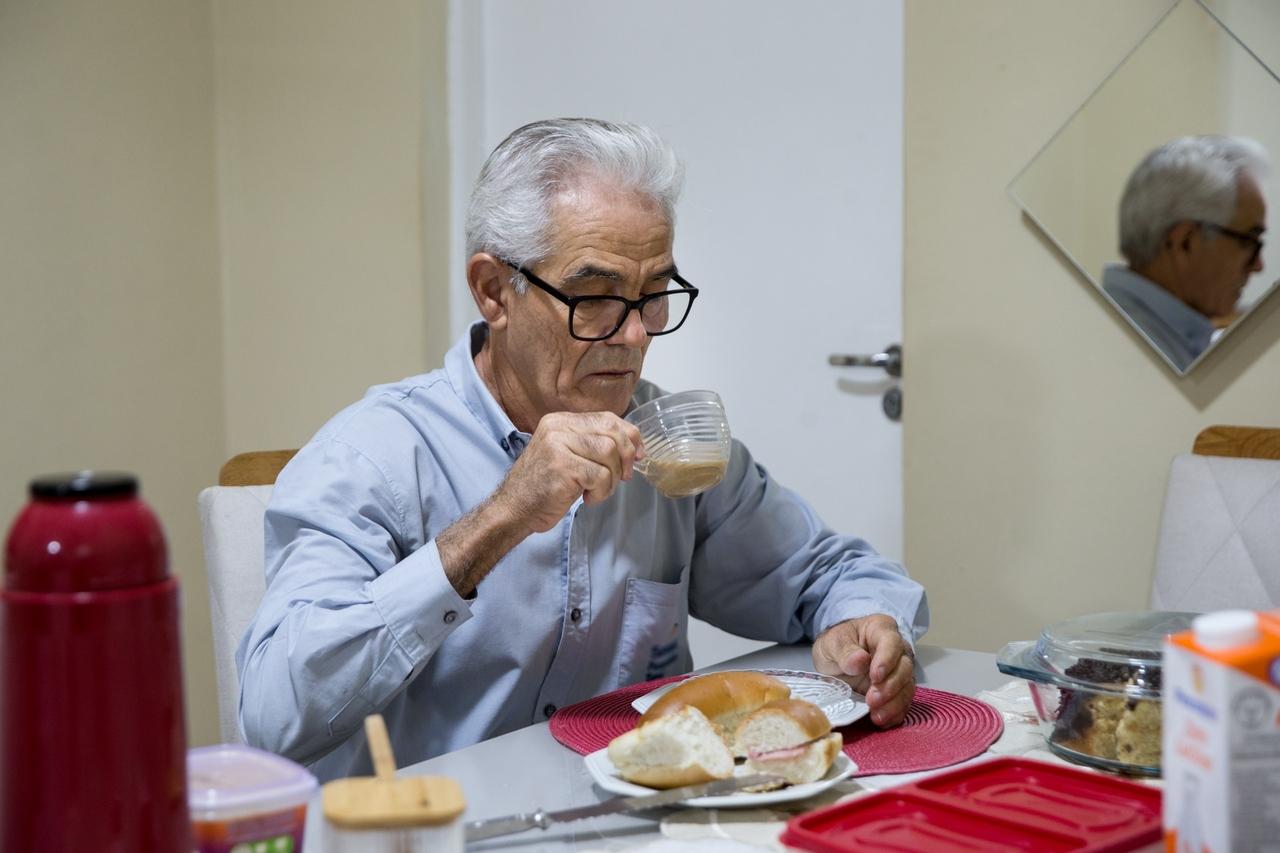 Homem idoso sentado na mesa de café da manhã, levando xícara de café com leite à boca.