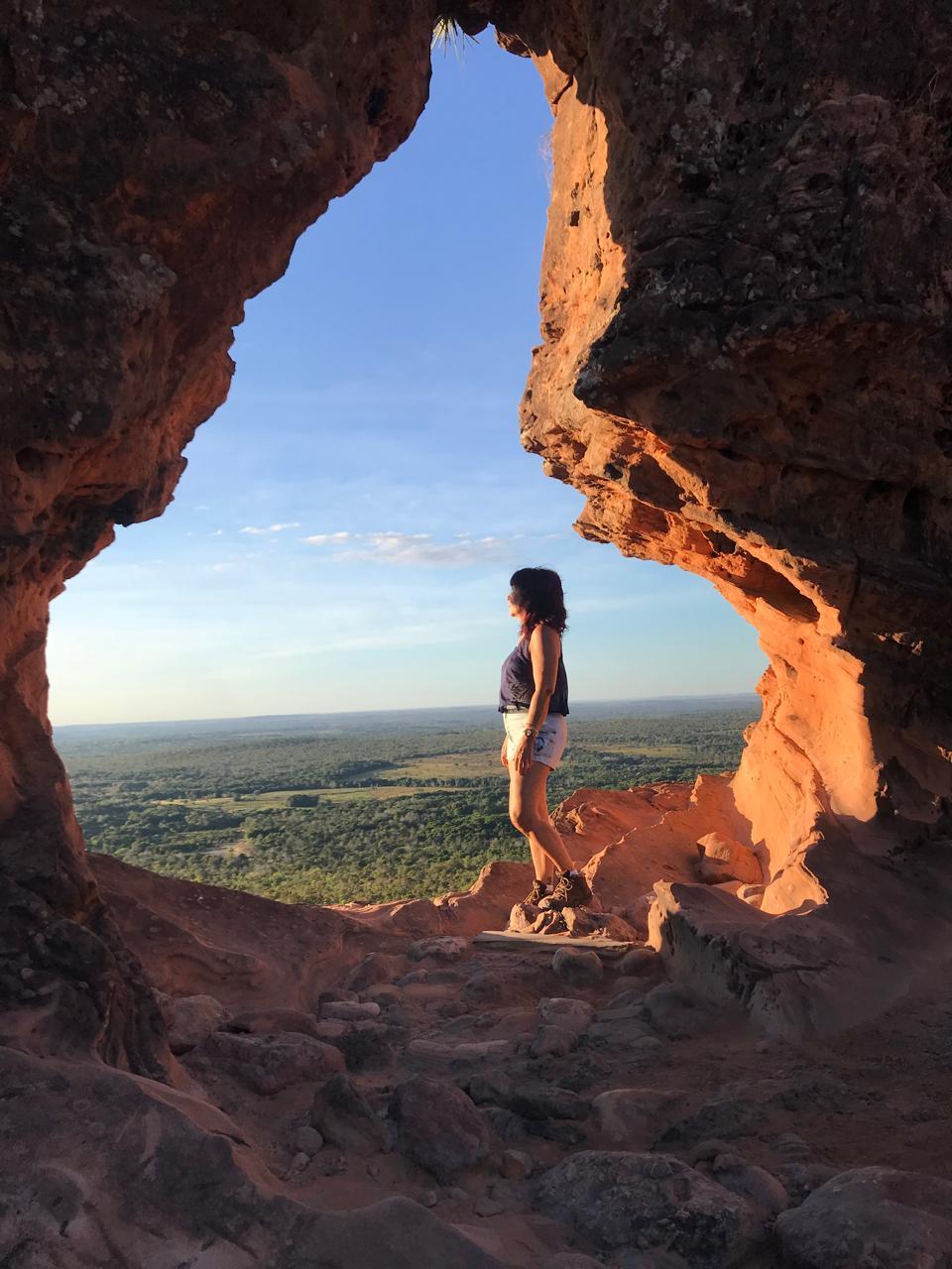 Mulher aparentemente no topo de uma montanha, emoldurada por uma formação rochosa, olhando para o horizonte em dia ensolarado.