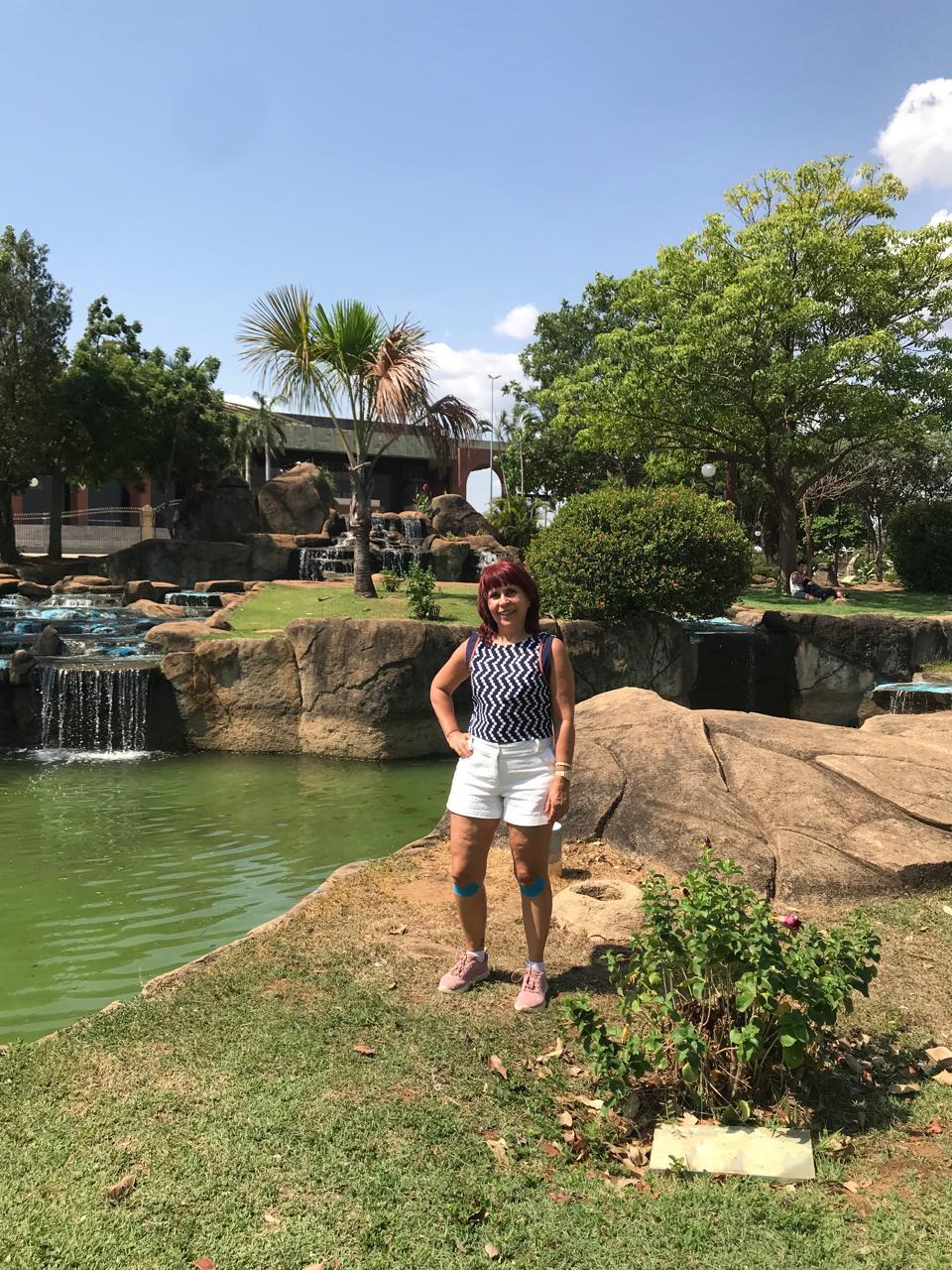 Mulher posa para foto em dia ensolarado em um local com grama verde e árvores.