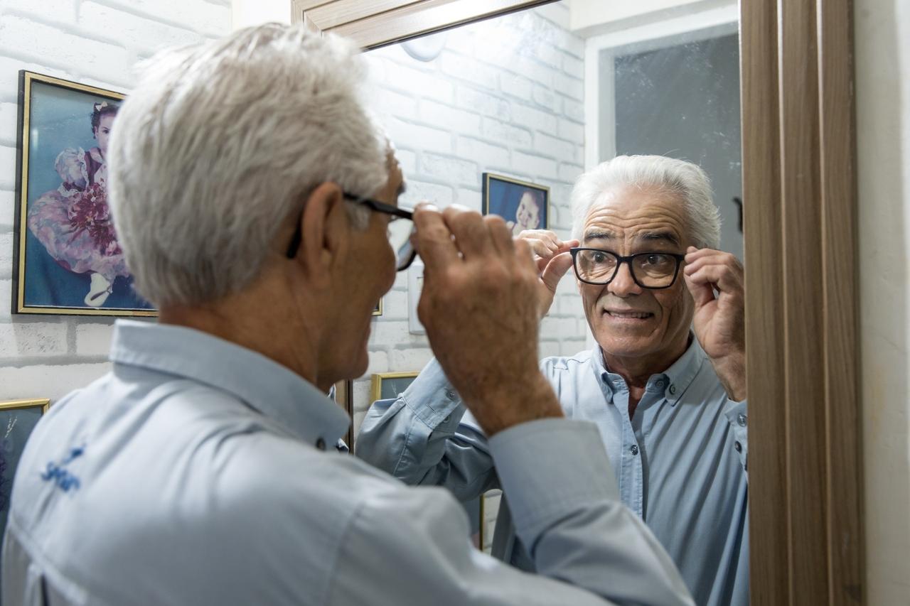 Homem idoso no espelho, arurmando os óculos de grau no rosto.