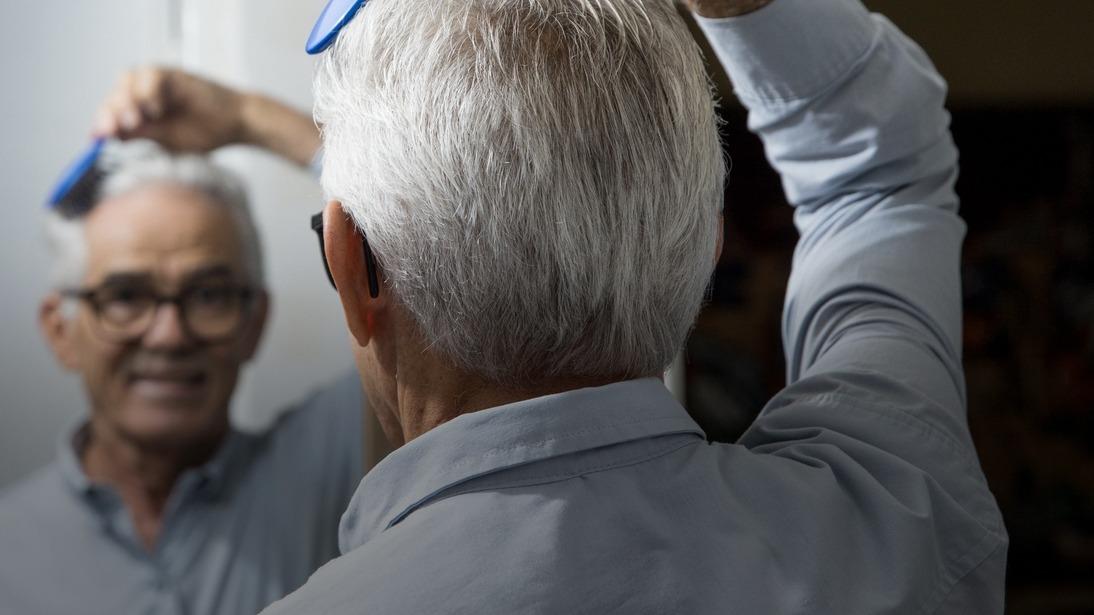 Idoso aparece de costas penteando o cabelo com uma escova azul, enquanto o seu reflexo aparece no espelho.