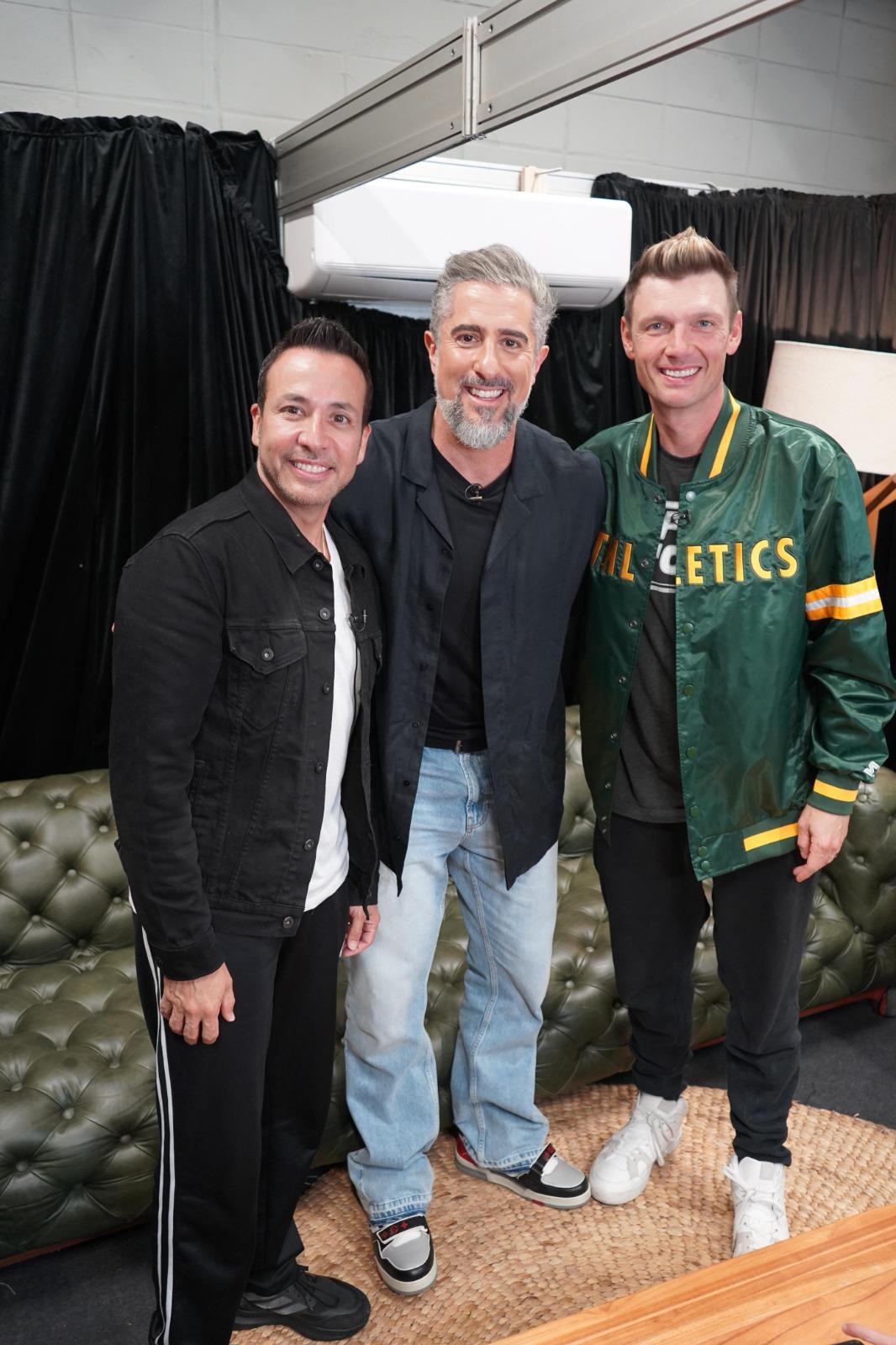 A foto mostra três homens sorrindo, posando lado a lado em um ambiente interno.