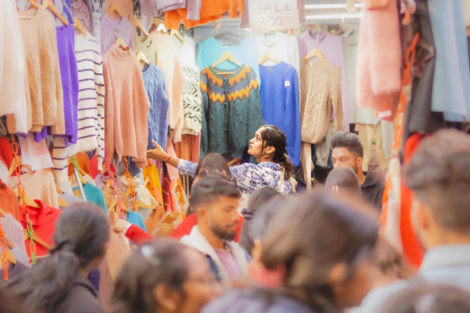 Mulher em um mercado movimentado observa suéteres coloridos pendurados em cabides, rodeada por várias pessoas entre as araras de roupas