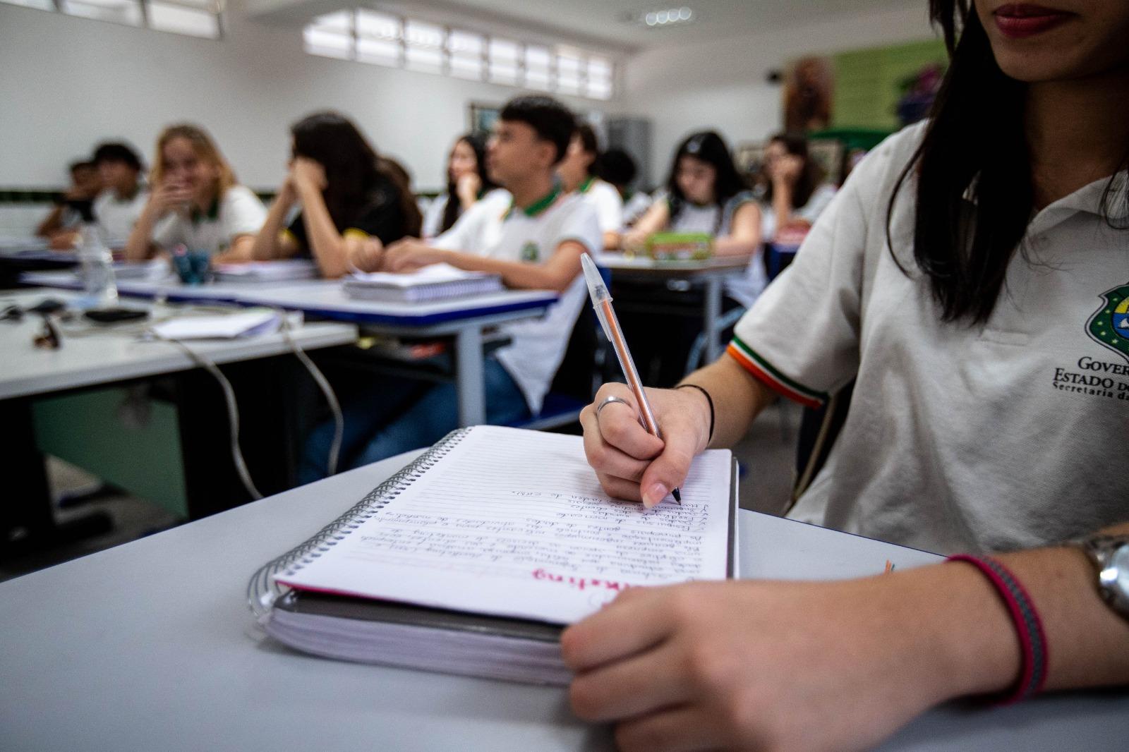Estudantes em sala de aula, com foco em uma aluna escrevendo no caderno enquanto colegas ao fundo acompanham a aula
