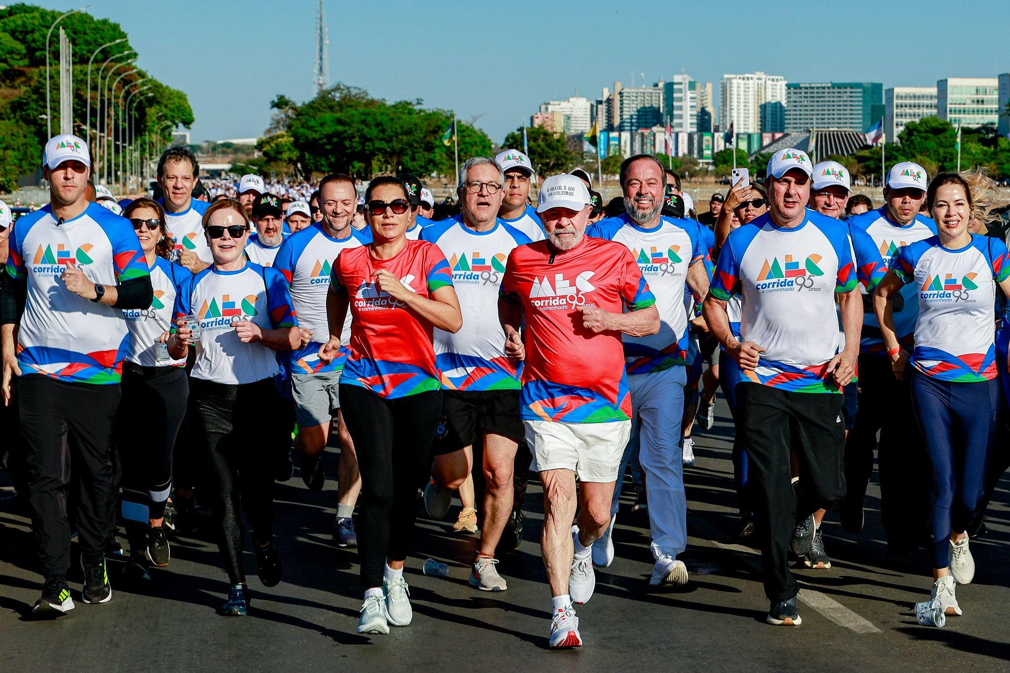 Grupo de pessoas participa de uma corrida ao ar livre em uma avenida larga e ensolarada. No centro da imagem, o presidente Lula e a primeira-dama Janta, ambos com camisetas vermelhas do evento, correm lado a lado e lideram o grupo. Ao redor deles, auxiliares do presidente vestem camisetas brancas e azuis com o mesmo logotipo da corrida. Ao fundo, aparecem árvores, bandeiras e prédios.