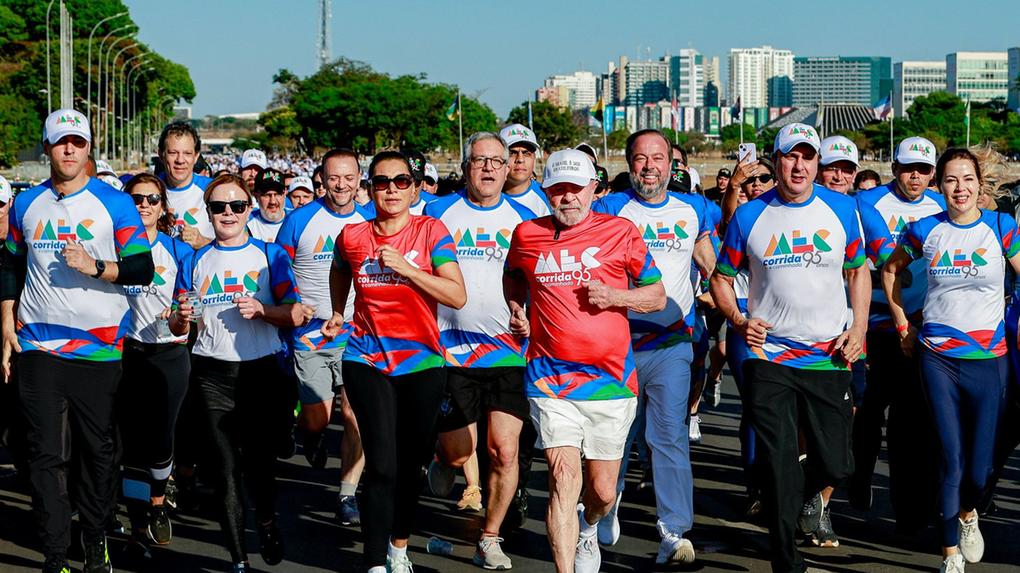 Grupo de pessoas participa de uma corrida ao ar livre em uma avenida larga e ensolarada. No centro da imagem, o presidente Lula e a primeira-dama Janta, ambos com camisetas vermelhas do evento, correm lado a lado e lideram o grupo. Ao redor deles, auxiliares do presidente vestem camisetas brancas e azuis com o mesmo logotipo da corrida. Ao fundo, aparecem árvores, bandeiras e prédios.