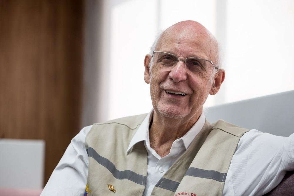 Na imagem, padre Júlio Lancellotti está sentado em sofá do Sistema Verdes Mares e sorri durante entrevista; ele traja o colete da Pastoral do Povo da Rua