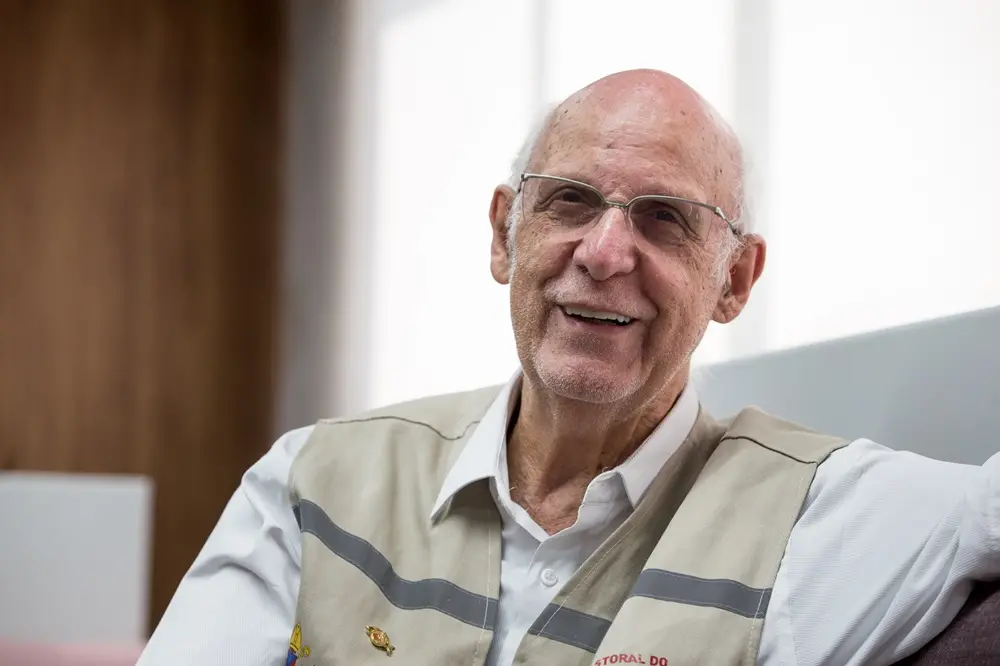 Na imagem, padre Júlio Lancellotti está sentado em sofá do Sistema Verdes Mares e sorri durante entrevista; ele traja o colete da Pastoral do Povo da Rua