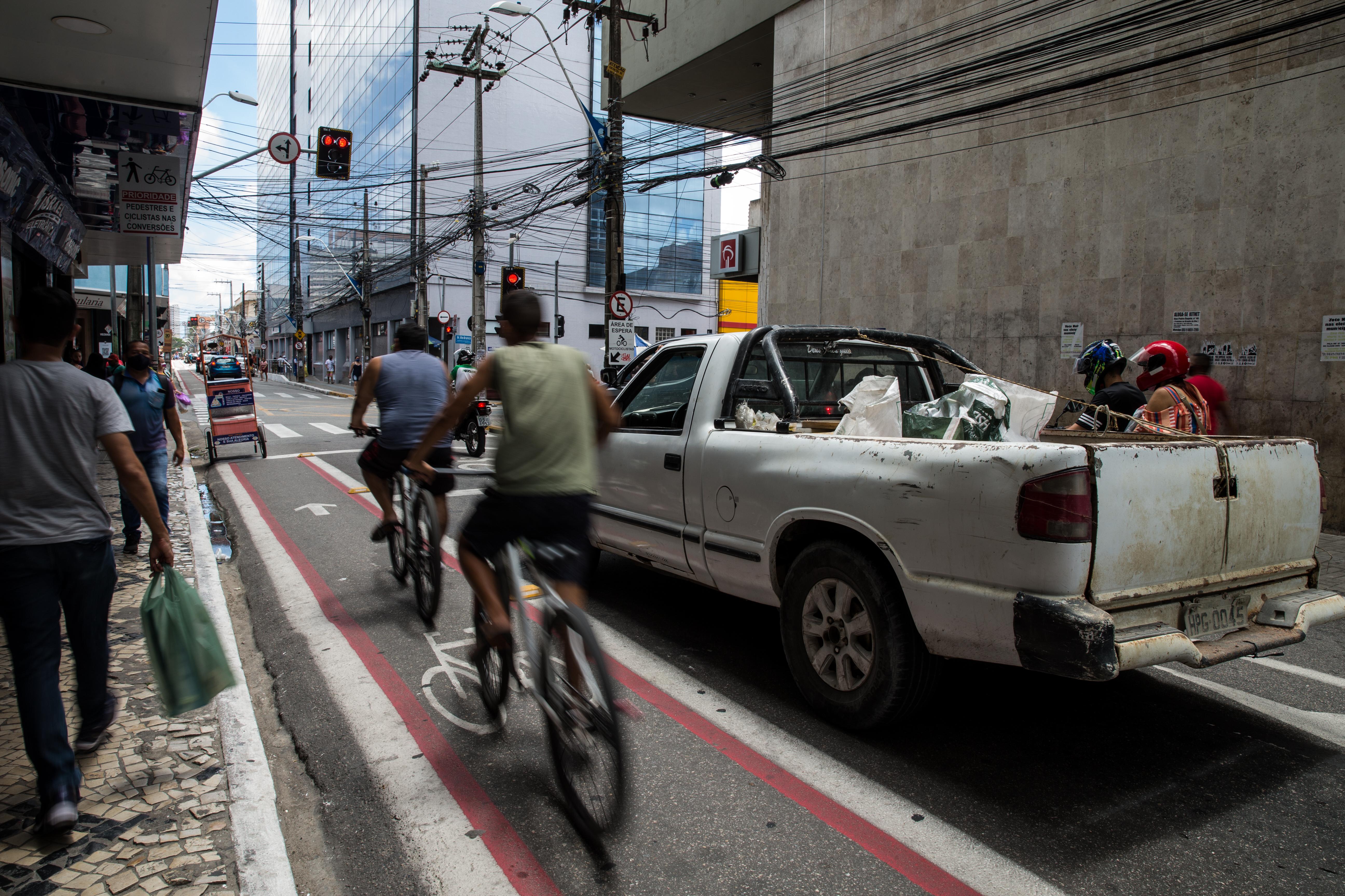 Vista da rua com duas pessoas em bicicletas, capturadas com desfoque de movimento, pedalando em uma faixa vermelha demarcada ao lado de uma picape branca suja. No fundo, há um prédio moderno de vidro, fiação elétrica aérea e pedestres na calçada.