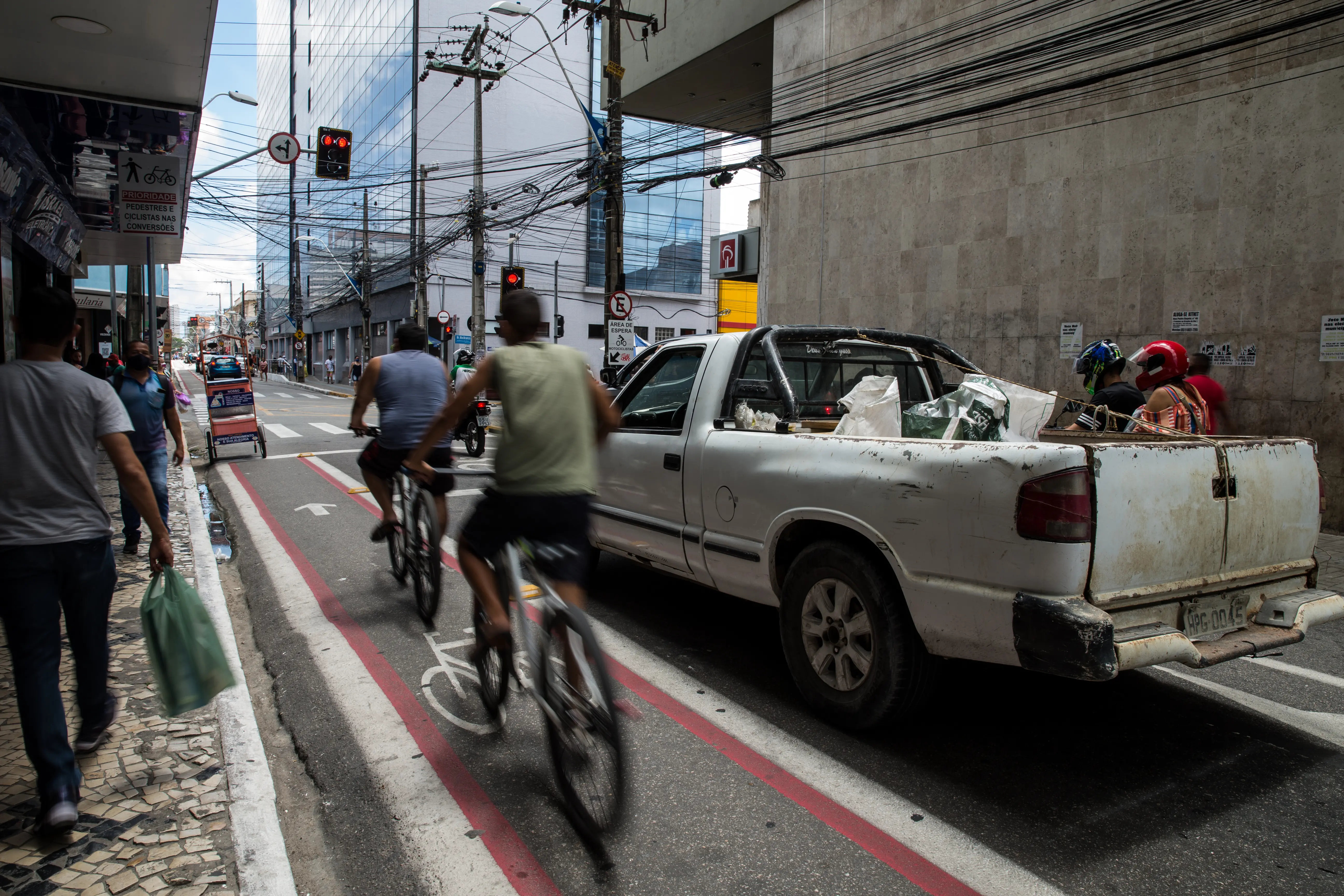 Vista da rua com duas pessoas em bicicletas, capturadas com desfoque de movimento, pedalando em uma faixa vermelha demarcada ao lado de uma picape branca suja. No fundo, há um prédio moderno de vidro, fiação elétrica aérea e pedestres na calçada.