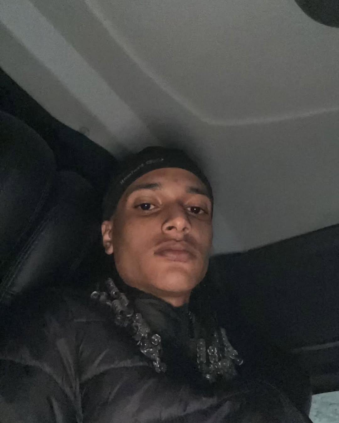 Imagem mostra rapper Oruam, com cabelo com tranças e expressão séria, usando jaqueta preta e beanie, sentado no interior de um carro escuro.