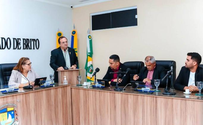 Reunião em plenário da câmara municipal de Barroquinha, com cinco pessoas sentadas em torno de uma mesa em formato de “U” de madeira clara. Um homem de terno escuro está em pé, falando em um púlpito ao centro. Atrás dele, três bandeiras estão posicionadas, incluindo a do Brasil e a do estado do Ceará. À mesa, quatro pessoas — duas mulheres e dois homens — acompanham a sessão, com microfones, copos de vidro e papéis à frente.