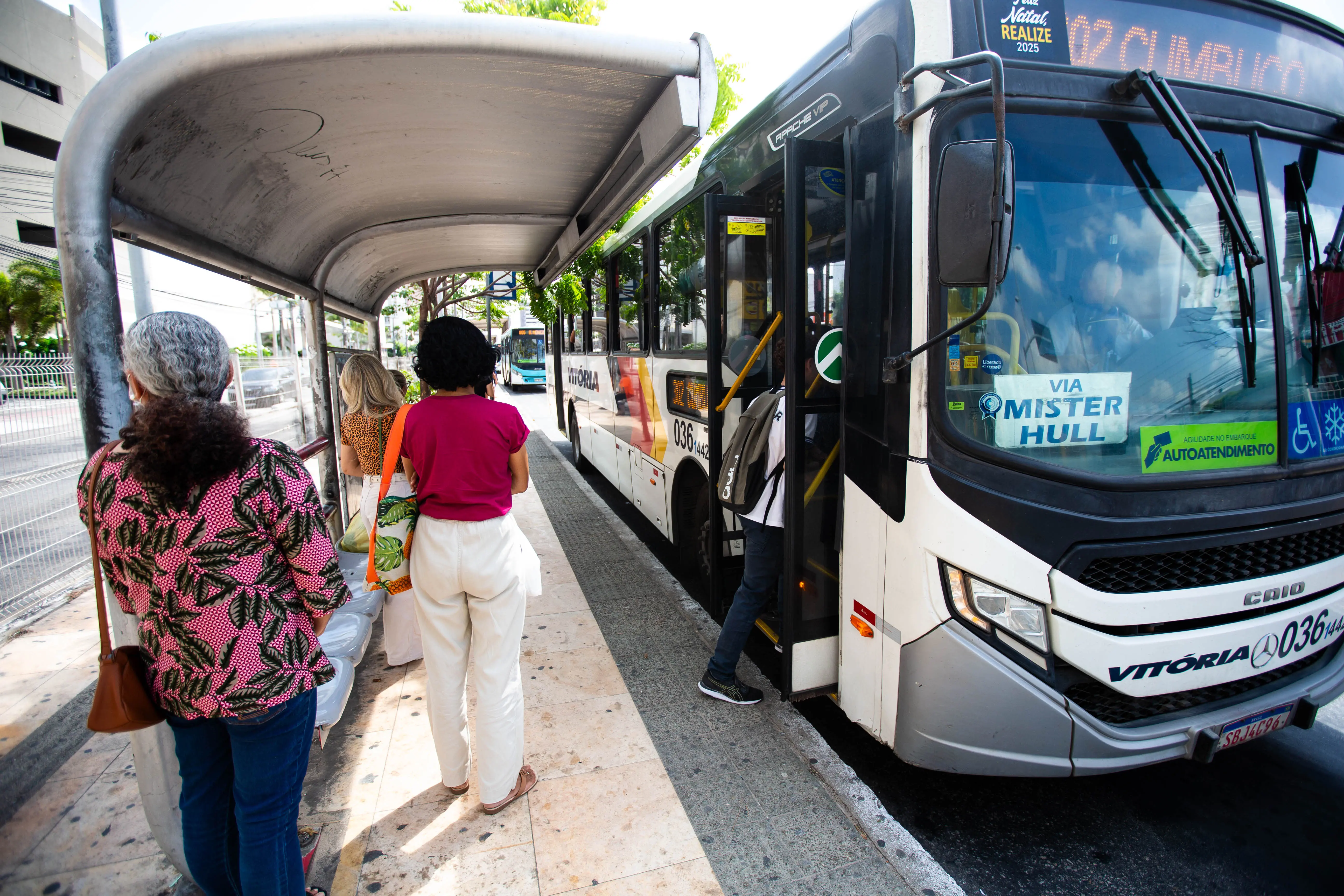 Uma fotografia em ângulo ligeiramente baixo e aberto captura um ônibus de transporte metropolitano parado em um ponto para embarque e desembarque.O ônibus, predominantemente branco com detalhes em preto e um adesivo colorido grande na lateral, ocupa o lado direito da imagem. No lado esquerdo, sob um abrigo de ponto de ônibus metálico, há três mulheres de costas esperando. A mulher em primeiro plano, à esquerda, tem cabelos grisalhos e usa uma blusa preta e rosa com estampa floral. A mulher no meio veste uma camiseta magenta e calças brancas. O ambiente geral é urbano, com alguma vegetação no fundo, à esquerda, e o foco é na interação entre os passageiros e o veículo de transporte público.