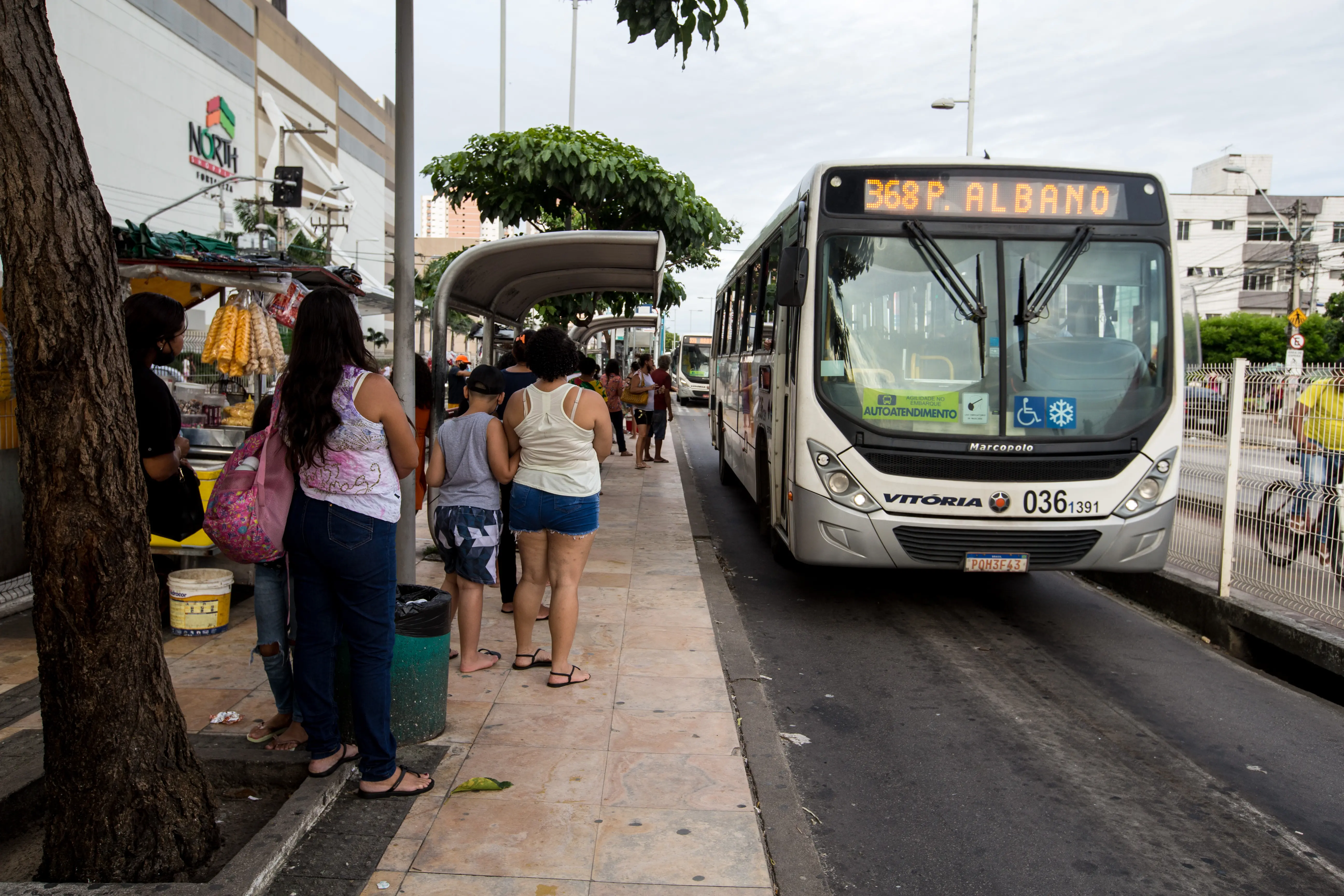 Ônibus de transporte público branco e cinza parado em um ponto de ônibus na rua. Várias pessoas estão esperando sob uma cobertura do ponto, e há uma árvore e uma fachada de loja visíveis no lado esquerdo.