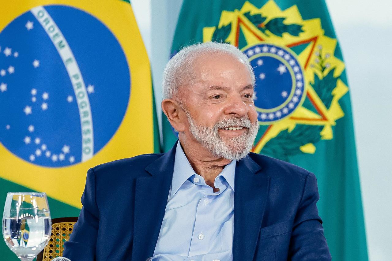 Imagem mostra presidente Lula sorrindo, usando terno azul e sem gravata, com bandeiras do Brasil ao fundo.