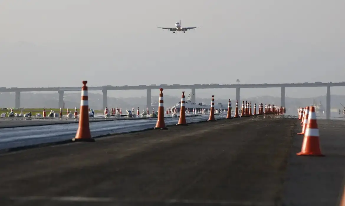 Imagem mostra avião decolando no Aeroporto Santos Dumont, no Rio de Janeiro, com cones de segurança na pista e uma ponte ao fundo, sob céu nublado.