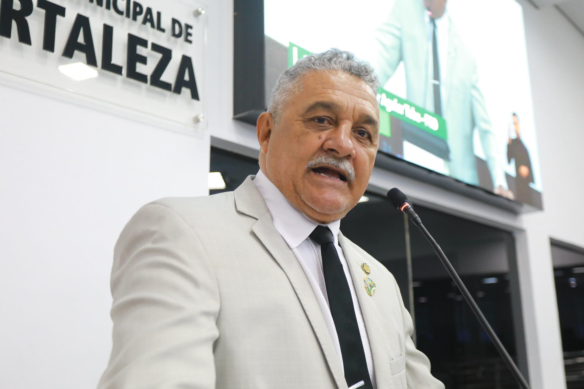 Professor Aguiar Toba discursa na tribuna da Câmara Municipal de Fortaleza. Ele é um homem idoso de cabelo branco e está de terno branco, gravata preta e camisa social branca.