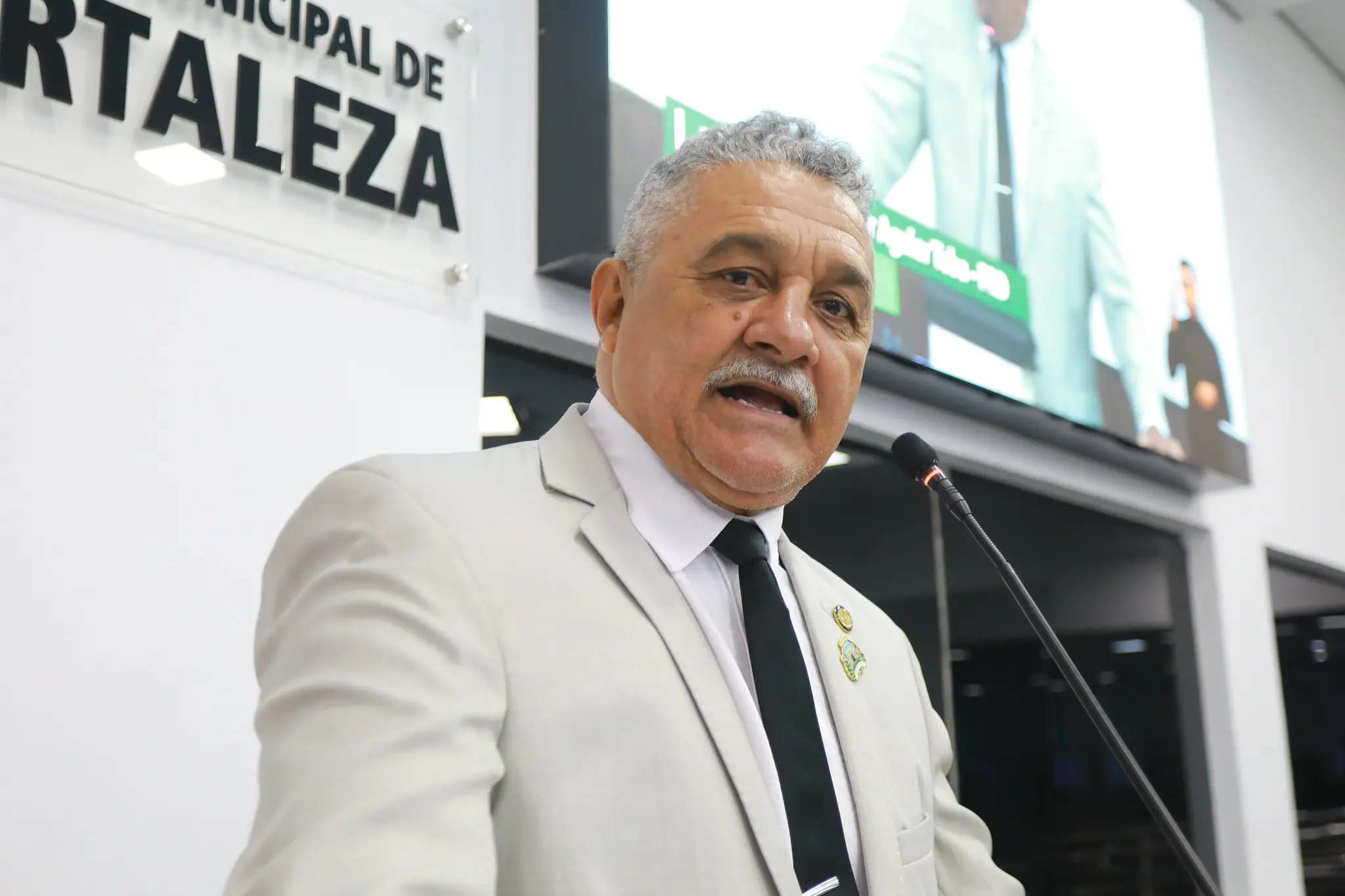 Professor Aguiar Toba discursa na tribuna da Câmara Municipal de Fortaleza. Ele é um homem idoso de cabelo branco e está de terno branco, gravata preta e camisa social branca.