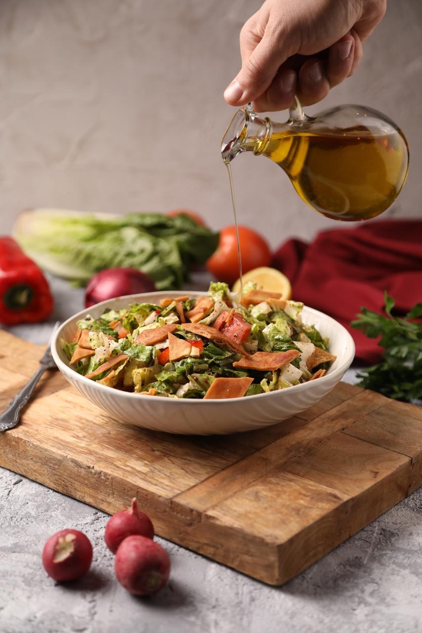 Salada fresca de alface, tomate e cenoura sendo regada com azeite de oliva extra virgem, em uma tábua de madeira, com ingredientes de legumes ao fundo.