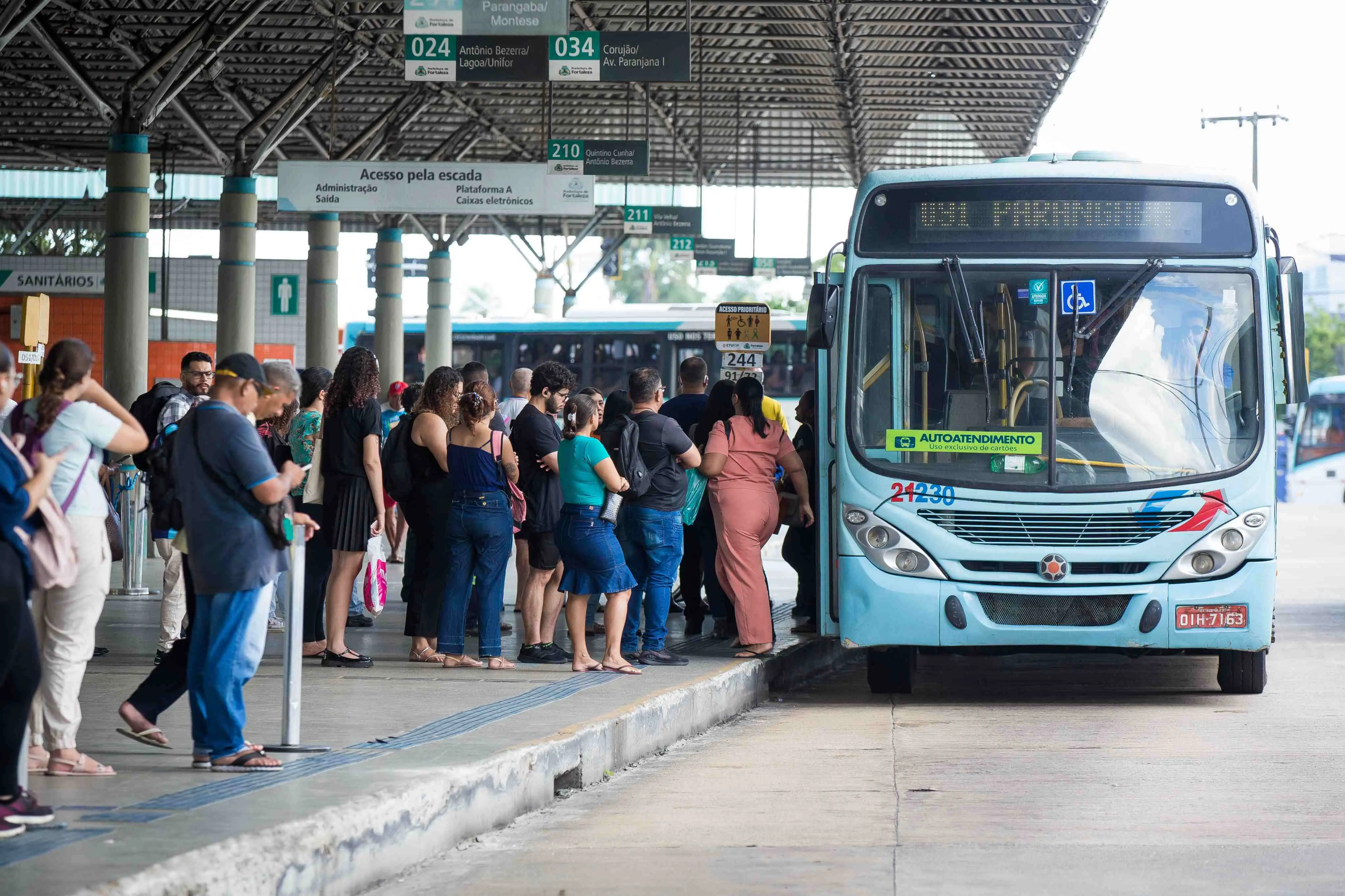 Uma foto em nível, tirada de perto, mostra uma fila de pessoas em uma plataforma de concreto em um terminal de ônibus. Um ônibus azul claro grande, moderno, com a porta aberta, está parado bem em frente à fila. As pessoas na fila são diversas e vestem roupas casuais. Elas estão voltadas para o ônibus. No fundo, há uma estrutura metálica coberta que forma o terminal, com placas brancas que listam os números e nomes das linhas.