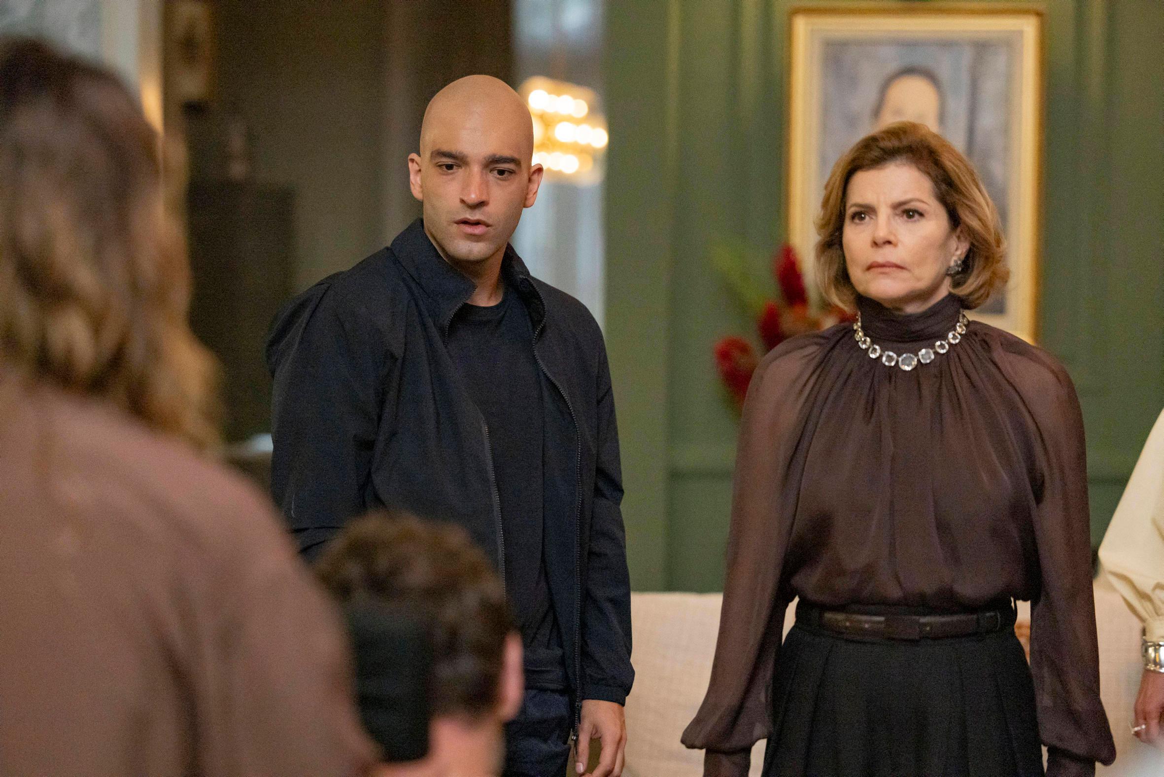 Cena da novela Vale Tudo mostra Afonso (Humberto Carrão), de aparência careca, vestindo camiseta e jaqueta preta, com expressão de surpresa. Ao lado dele está Odete Roitman (Debora Bloch), mulher mais velha, de cabelo curto castanho, usando blusa marrom elegante com colar de pedras, olhando séria e abalada. A cena acontece em um ambiente sofisticado, com parede verde, flores vermelhas e quadro ao fundo.