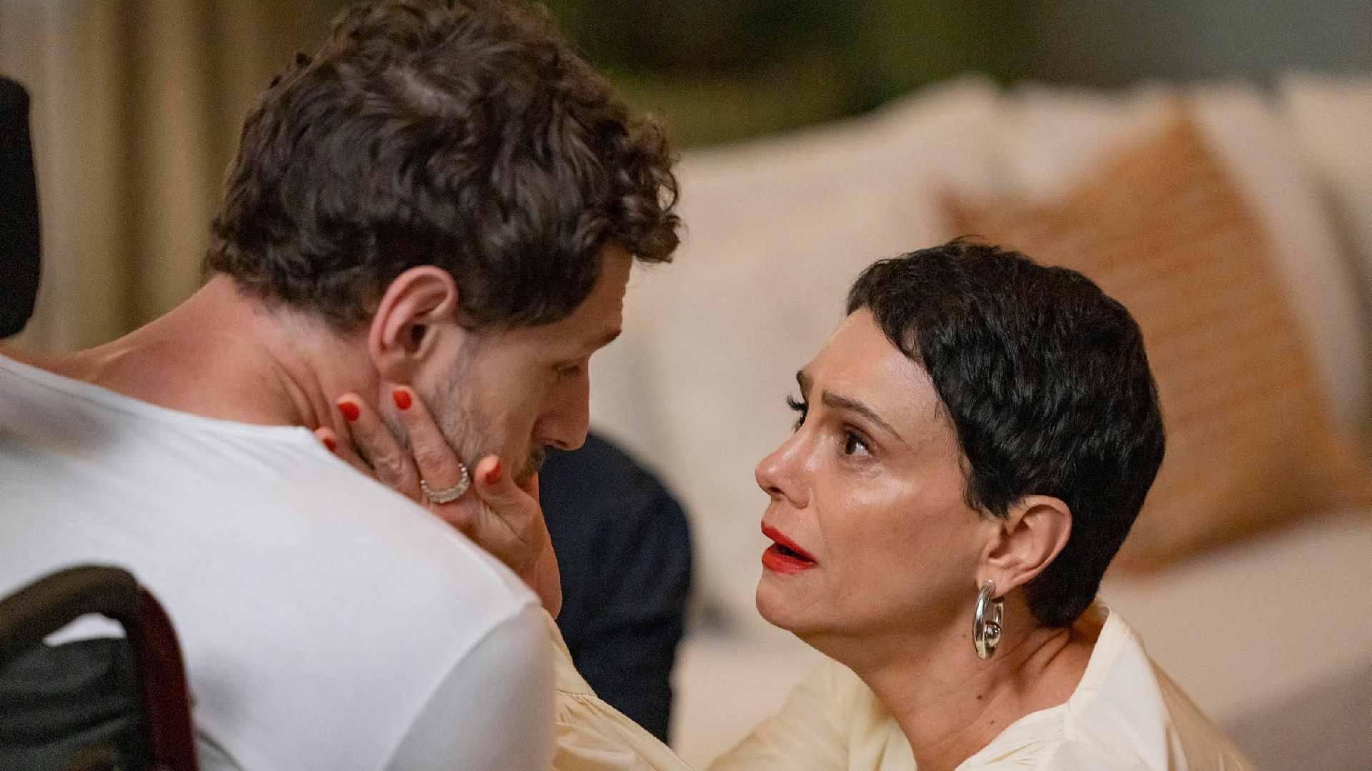 Cena da novela Vale Tudo mostra Celina, de cabelo curto e escuro, usando blusa clara e brincos grandes, olhando com emoção e espanto para o sobrinho Leonardo. Ela segura o rosto dele com as duas mãos enquanto fala com intensidade. Leonardo aparece de perfil, com expressão séria e cabisbaixa. A cena transmite tensão e surpresa em um ambiente de sala com sofá ao fundo.