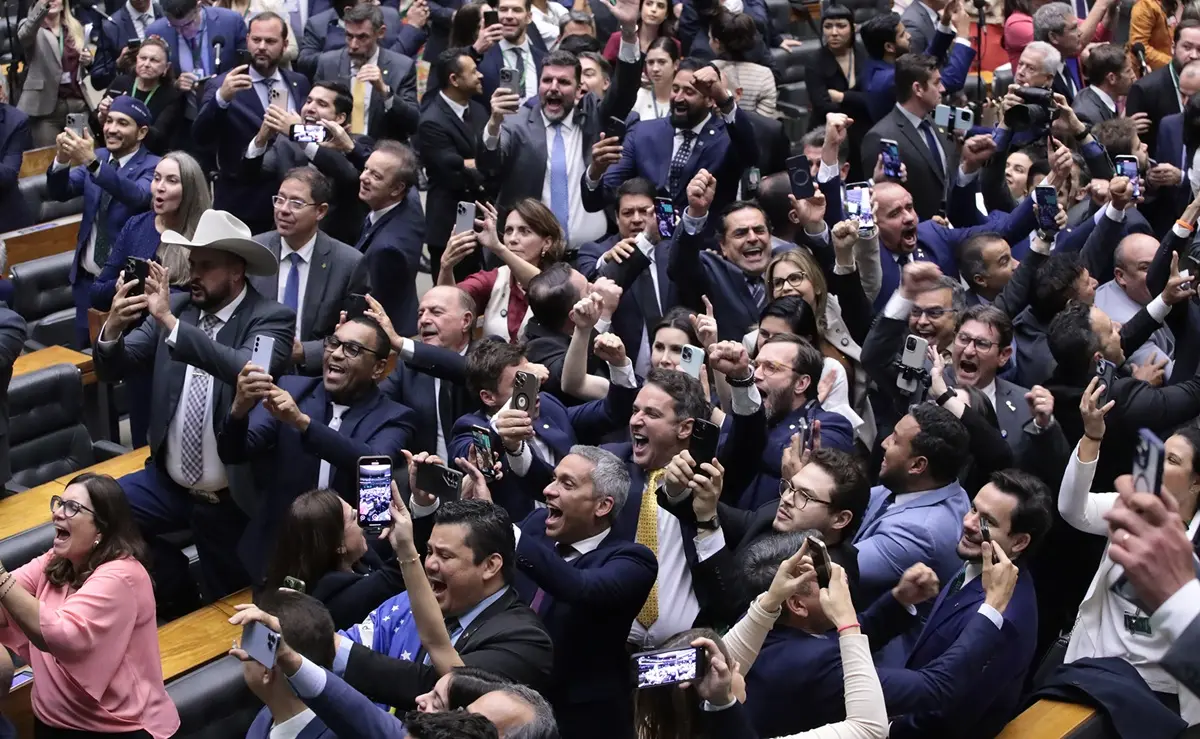 Grupo grande de deputados e deputadas em plenário comemora animadamente, muitos com celulares em mãos registrando o momento de aprovação da urgência do PL da Anistia, alguns com braços erguidos e gritos de euforia.