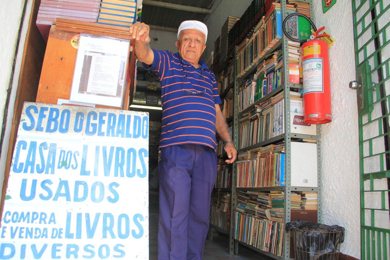 Imagem mostra Geraldo Paulo Duarte, fundador do sebo O Geraldo, um dos mais tradicionais do Centro de Fortaleza, em 2014. Com expressão série e vestindo blusa azul escura listrada de laranja, calça também em azul-escuro e boina branca, ele posa ao lado no porta de entrada do sebo ao lado de uma placa que diz sebo do geraldo.