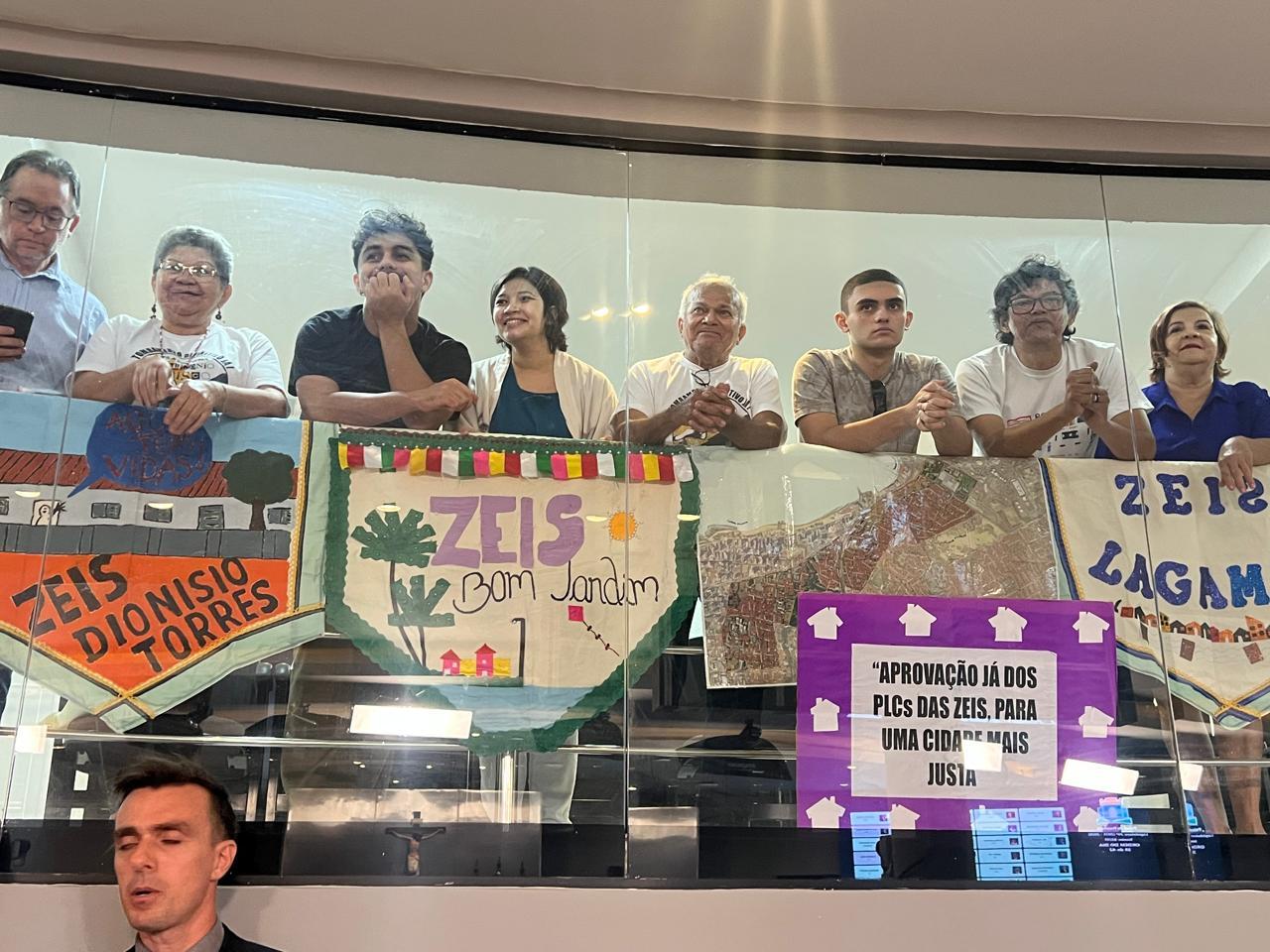 Grupo de pessoas em uma galeria de vidro segurando faixas coloridas em defesa das ZEIS (Zonas Especiais de Interesse Social). Entre as faixas estão escritas: 'ZEIS Dionísio Torres', 'ZEIS Bom Jardim', 'ZEIS Lagamar' e um cartaz roxo com a frase: 'Aprovação já dos PLCs das ZEIS, para uma cidade mais justa'.