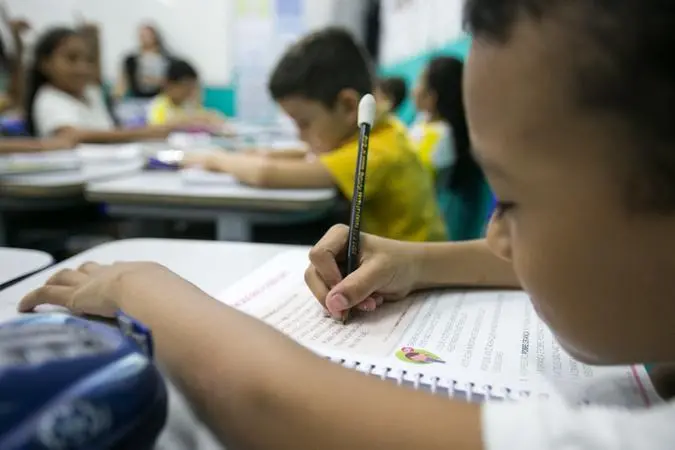 Menino escrevendo com um lápis no livro didático no ambiente de sala de aula.