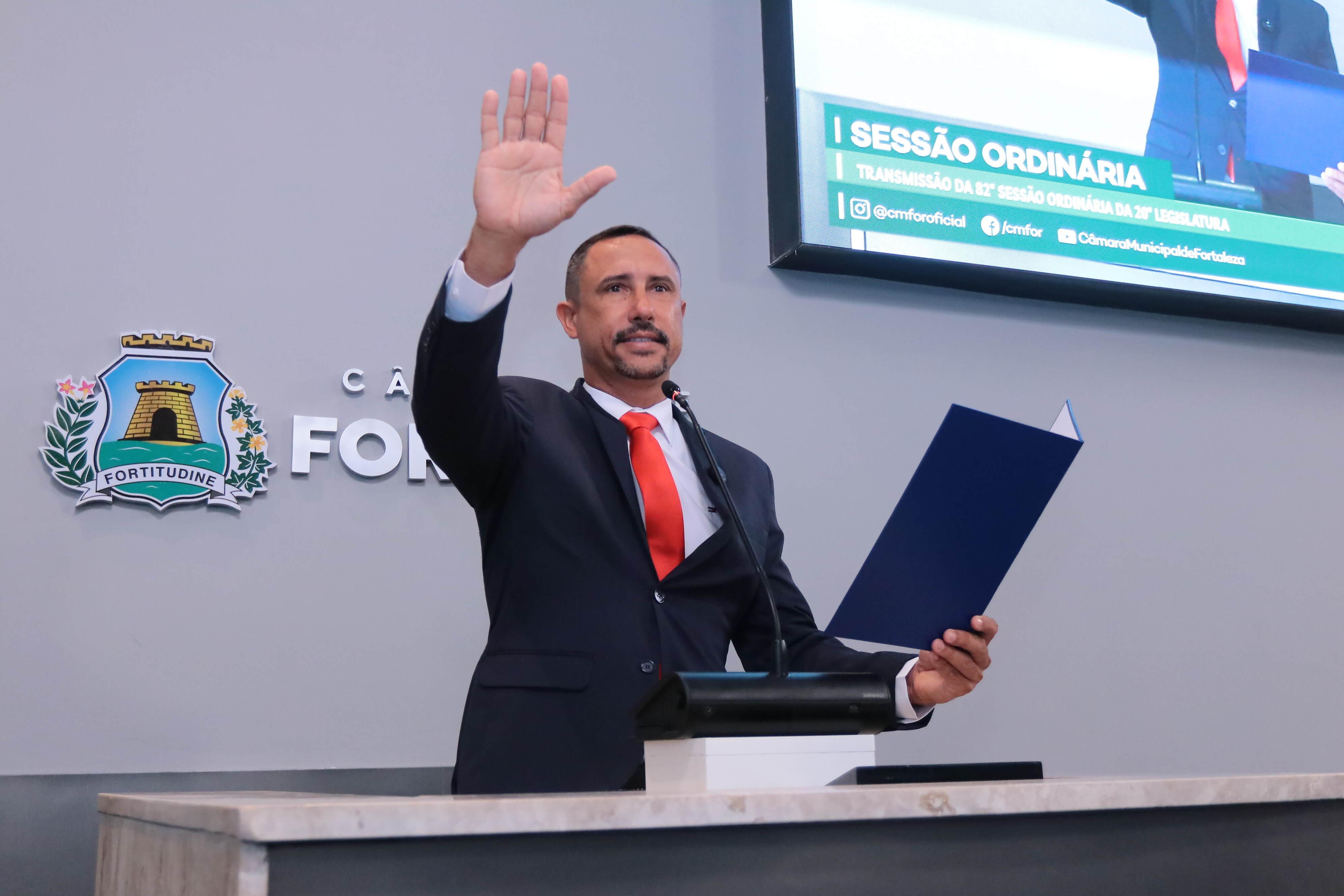 Imagem mostra um homem em pé atrás de um púlpito, em ambiente oficial da Câmara Municipal de Fortaleza. Ele veste terno escuro, camisa branca e gravata vermelha, segurando uma pasta azul em uma das mãos e levantando a outra mão direita, em gesto de juramento. Atrás dele, na parede, está o brasão da cidade de Fortaleza com a palavra “Fortitudine”. No canto superior direito, há um telão transmitindo a sessão ordinária da legislatura.
