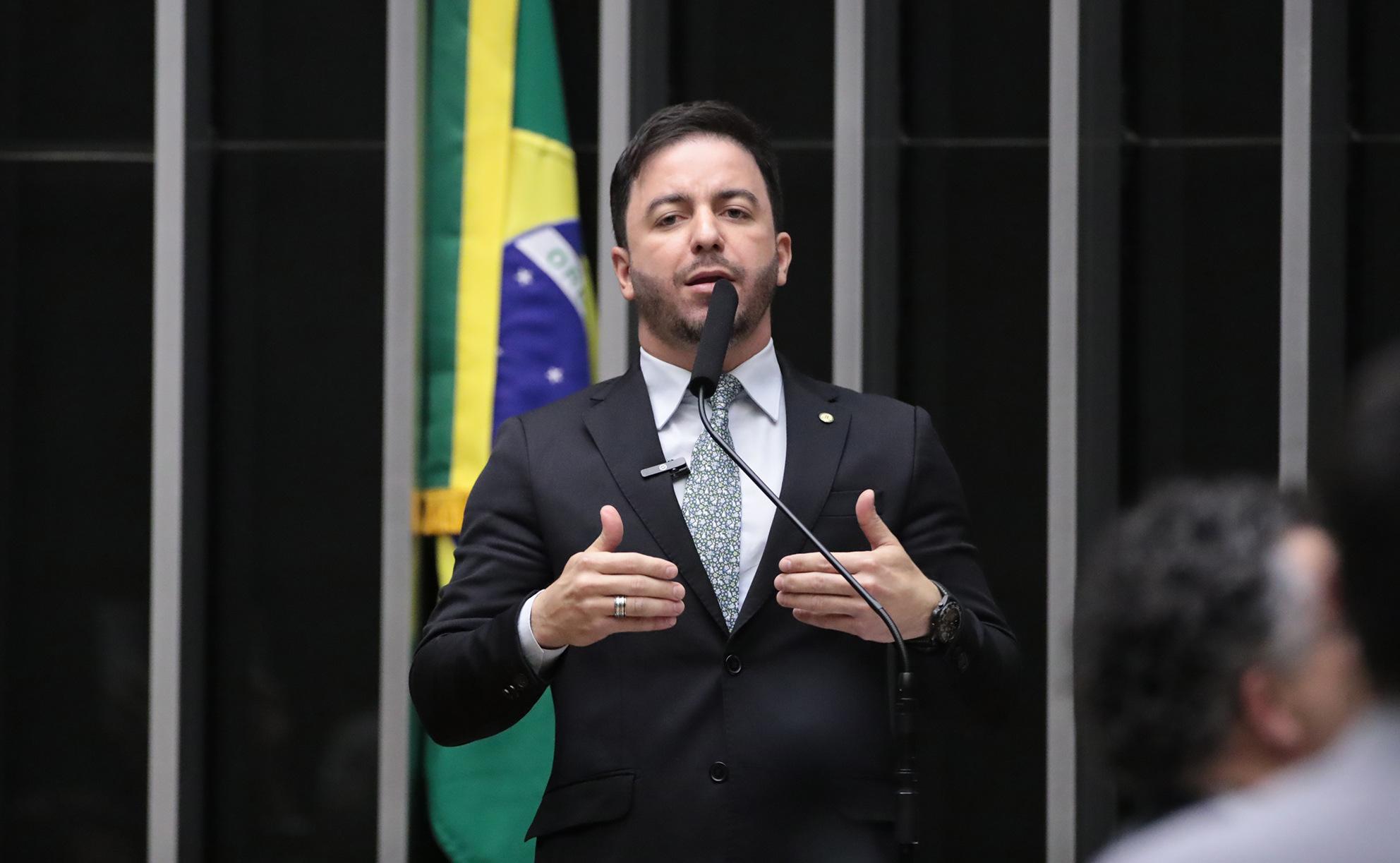 Imagem de um homem em pé, vestindo terno preto, camisa branca e gravata estampada em tons claros, falando ao microfone em um púlpito. Trata-se de Célio Studart. Ele gesticula com as mãos enquanto discursa. Ao fundo, aparece parcialmente a bandeira do Brasil.