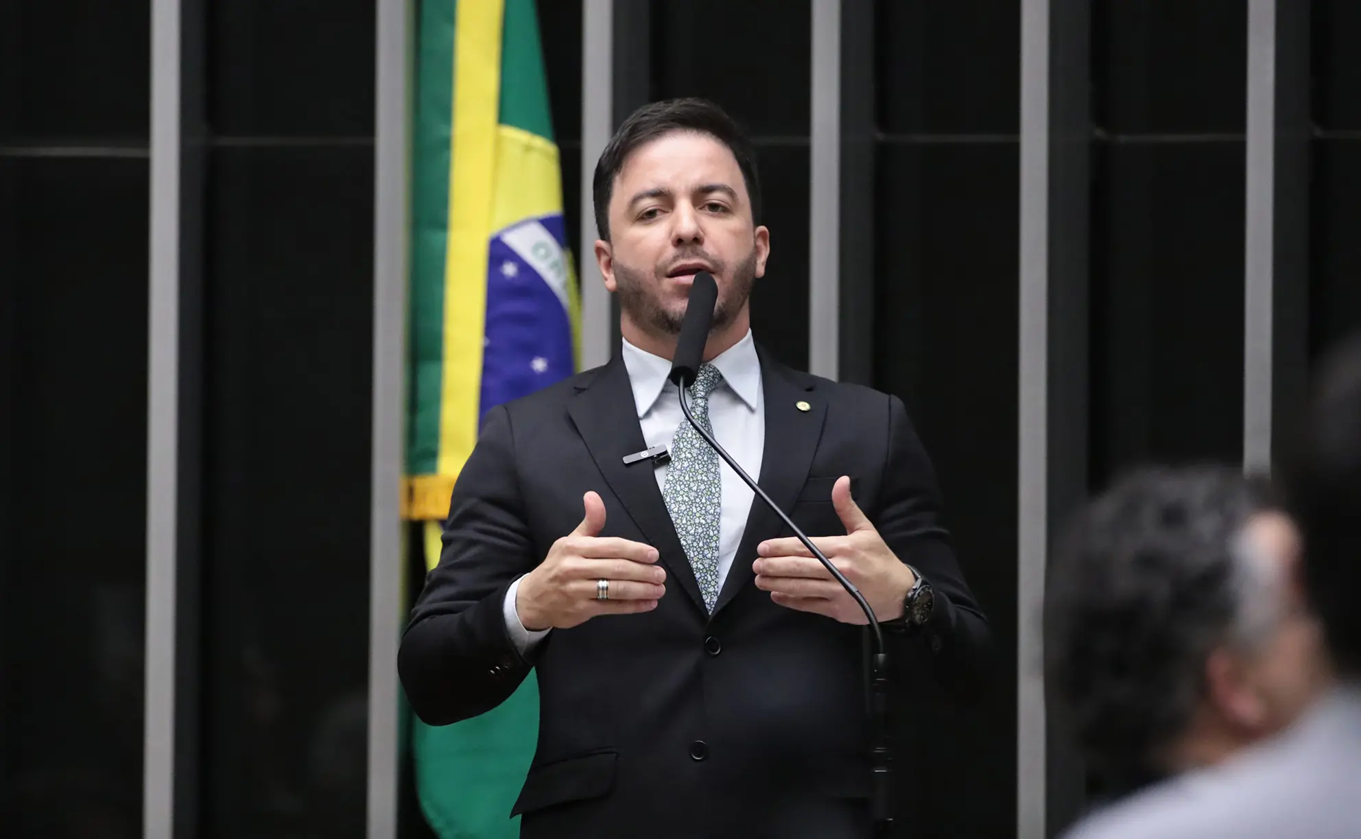 Imagem de um homem em pé, vestindo terno preto, camisa branca e gravata estampada em tons claros, falando ao microfone em um púlpito. Trata-se de Célio Studart. Ele gesticula com as mãos enquanto discursa. Ao fundo, aparece parcialmente a bandeira do Brasil.
