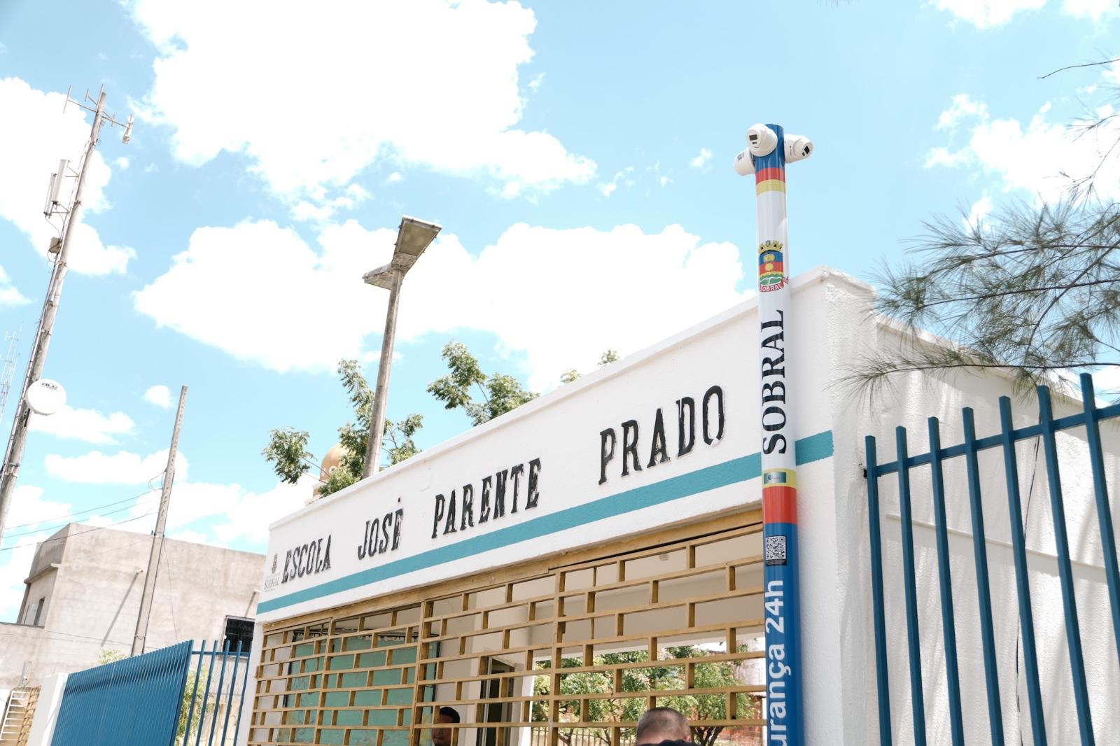 Fachada da Escola José Parente Prado, em Sobral, com portão metálico azul e poste de videomonitoramento de segurança 24h instalado na entrada