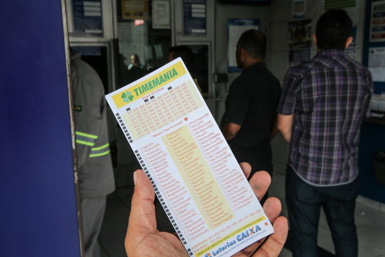 Pessoa segurando um bilhete de loteria da Timemania em uma agência lotérica, com várias pessoas aguardando na fila ao fundo. A imagem destaca a aposta na loteria.