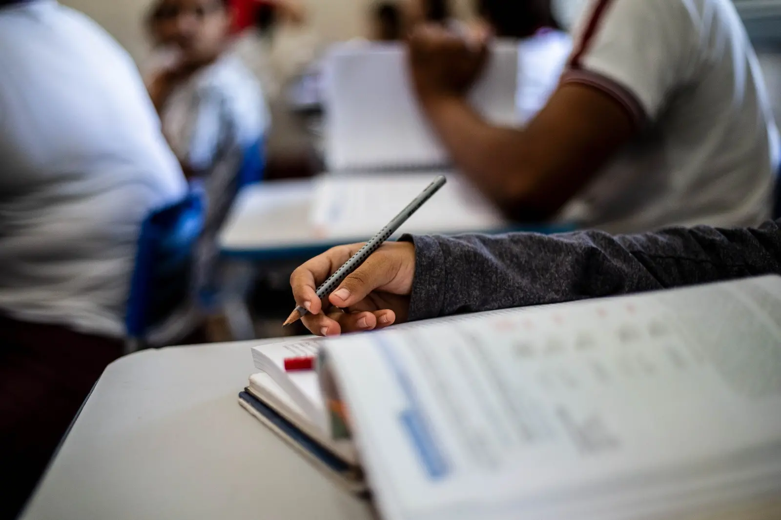 Mão de um estudante segurando um lápis sobre um livro, com outros alunos ao fundo em uma sala de aula.