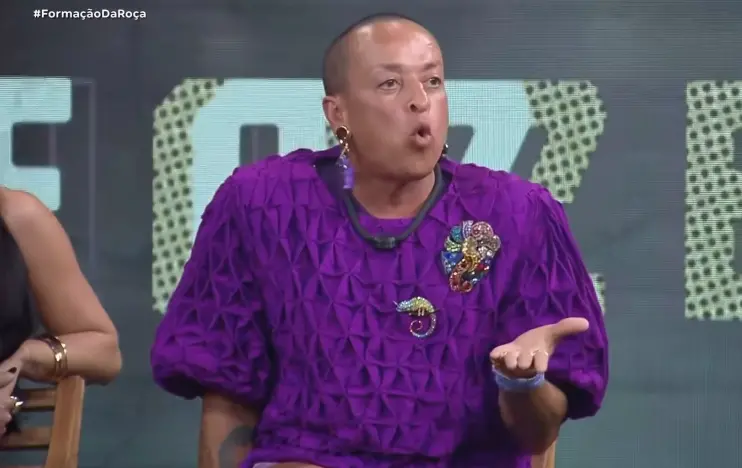 Homem com cabelo raspado e roupa roxa, fazendo uma expressão facial durante um debate, com fundo de estúdio e texto em destaque.