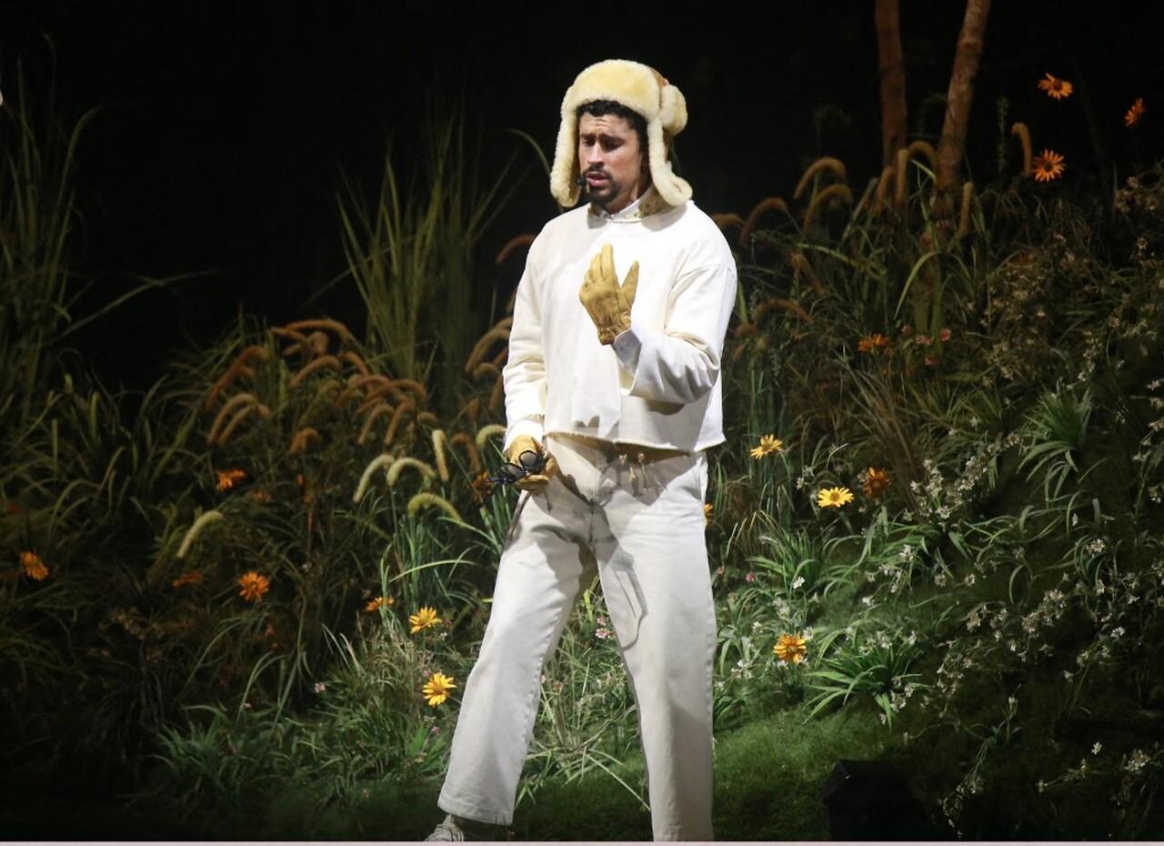 Cantor, vestido com roupas brancas e gorro de lã, atuando em palco ao ar livre com vegetação ao fundo.