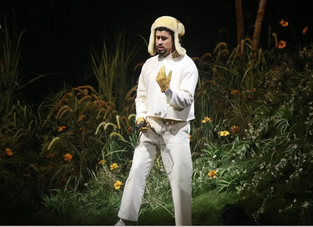 Cantor, vestido com roupas brancas e gorro de lã, atuando em palco ao ar livre com vegetação ao fundo.