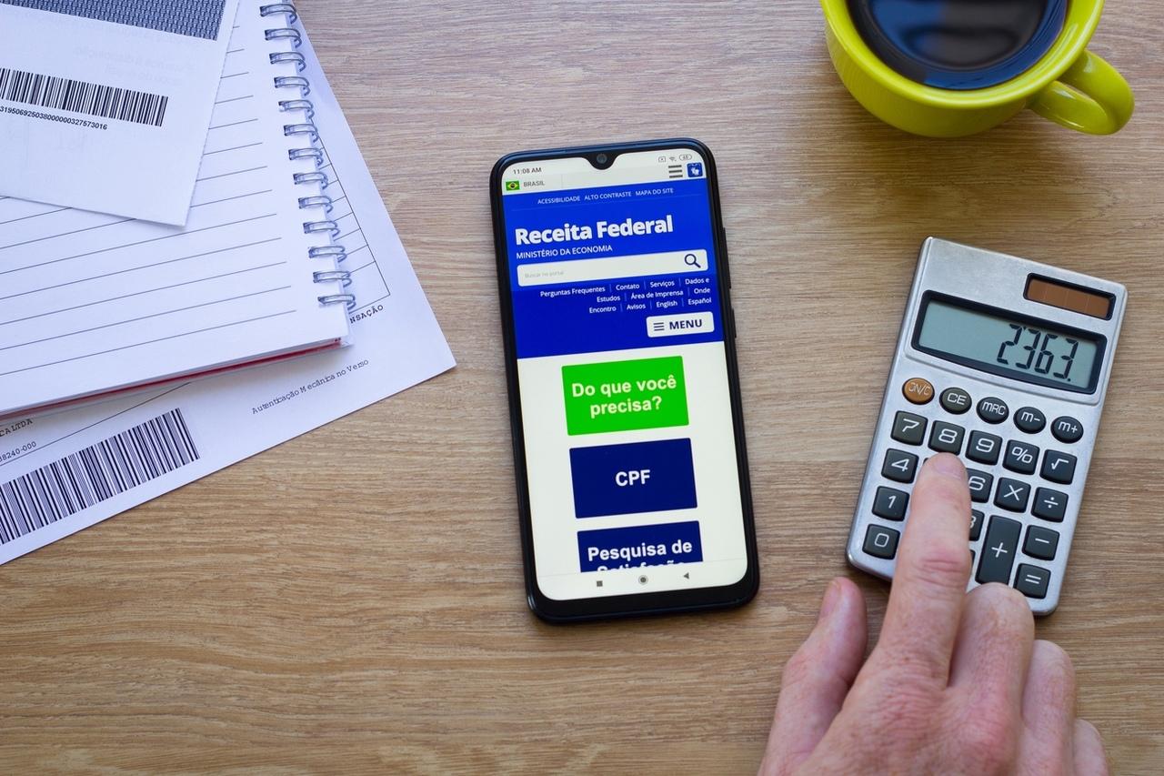 Imagem mostra celular com a tela exibindo a página da Receita Federal do Brasil. Esse celular está sobre uma mesa, ao lado de boletos à esquerda. À direita, uma calculadora aparece enquanto uma mão no canto da imagem digita os números 2,3,6 e 3. Acima, quase saindo do enquadramento, há uma xícara amarela com café.
