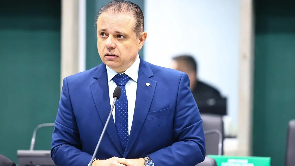 Homem de terno azul e gravata azul de bolinhas fala em um microfone em ambiente formal, possivelmente uma sessão parlamentar.